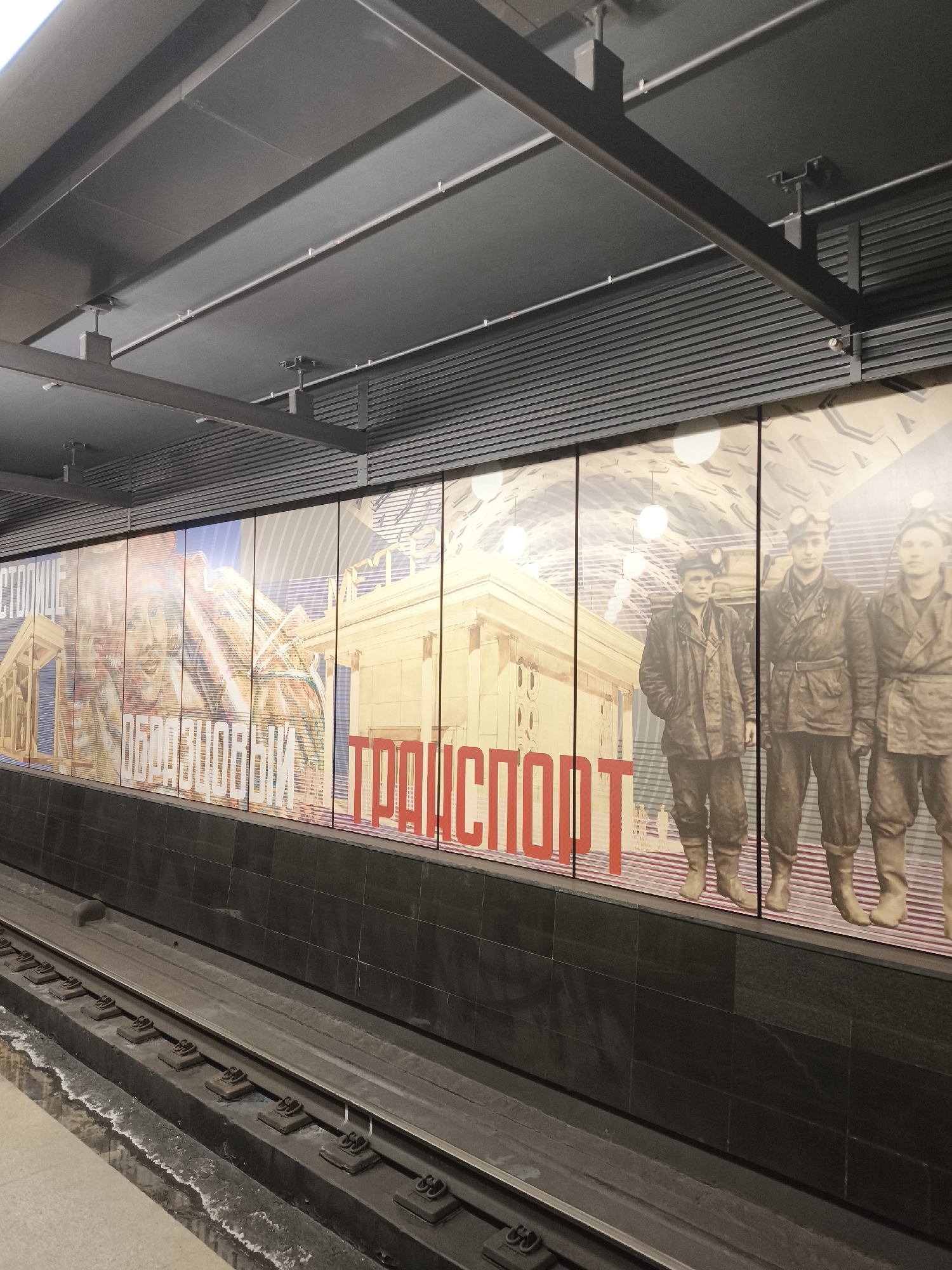 A wide-angle shot depicts a subway station platform. On the left is a portion of the platform floor, light grey in color. A set of train tracks runs along the lower-middle section of the image, with the ties visible between the rails. To the right of the tracks, a dark grey wall rises vertically.
Above the wall, a large rectangular panel, split into sections by vertical black lines, spans the length of the image. The panel displays a series of images and text in a style reminiscent of Soviet-era propaganda. On the left, there's a depiction of a building and a portrait. The word "ОБРАЗЦОВЫЙ" (Exemplary) is written in white, and the word "ТРАНСПОРТ" (Transport) is in red. To the right are figures of three people in working clothes, with headlamps, possibly miners.
The ceiling above the panel is grey and constructed of multiple rectangular panels. Metal beams support the ceiling structure. Overall, the lighting is even, and the scene appears to be indoors. The location is a subway station platform, in Unknown, Unknown.