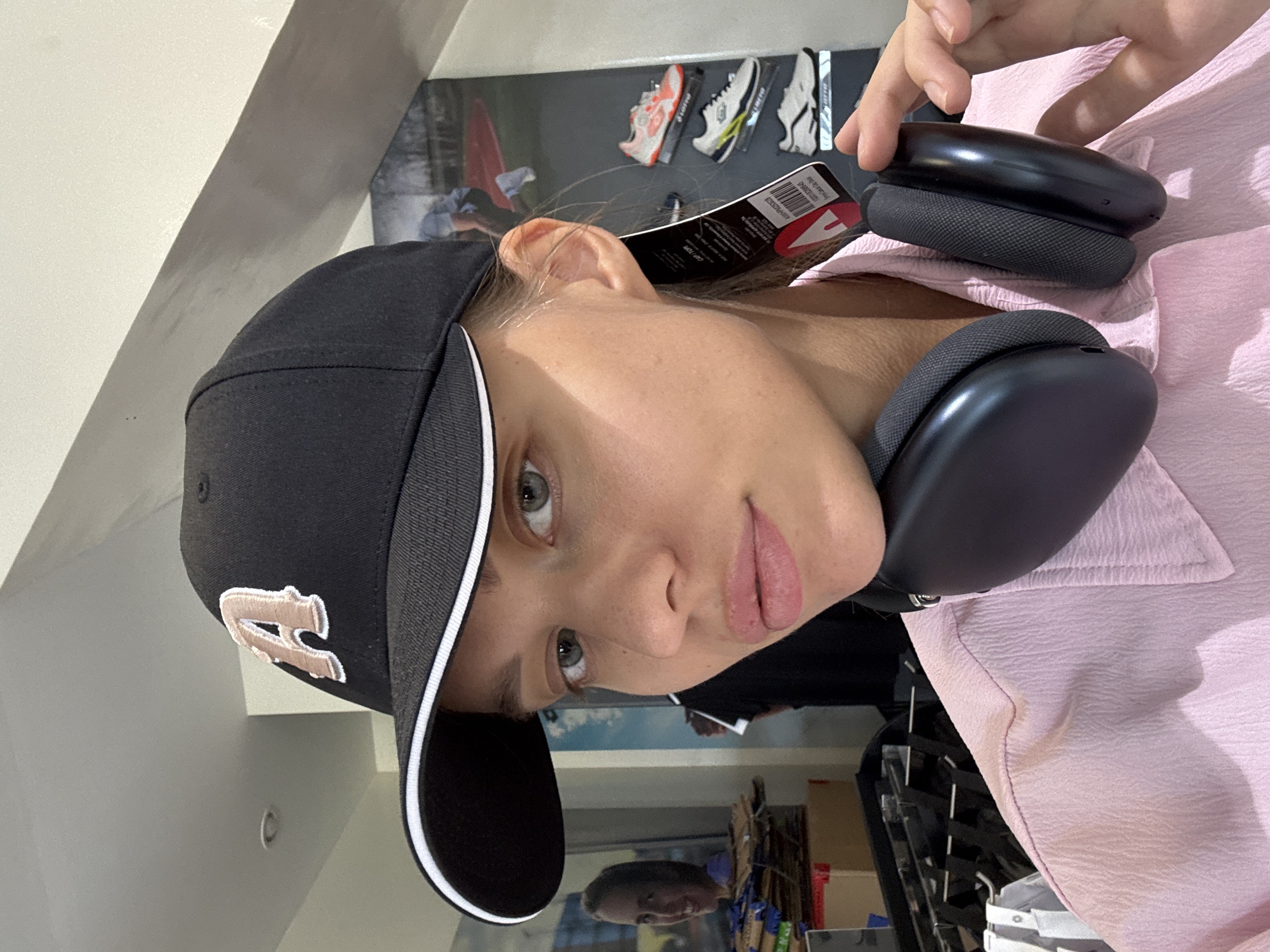 The image shows a woman wearing a black baseball cap with a stylized "A" on the front and white trim on the brim. She is also wearing black headphones with a price tag still attached. She has light skin, green eyes, and pink lips. She is wearing a pink collared shirt. The background appears to be a store, with shoes displayed on a wall. The lower part of the image shows an aisle in the store. There is a reflection of someone in the glass. The location is North Kuta, Indonesia.