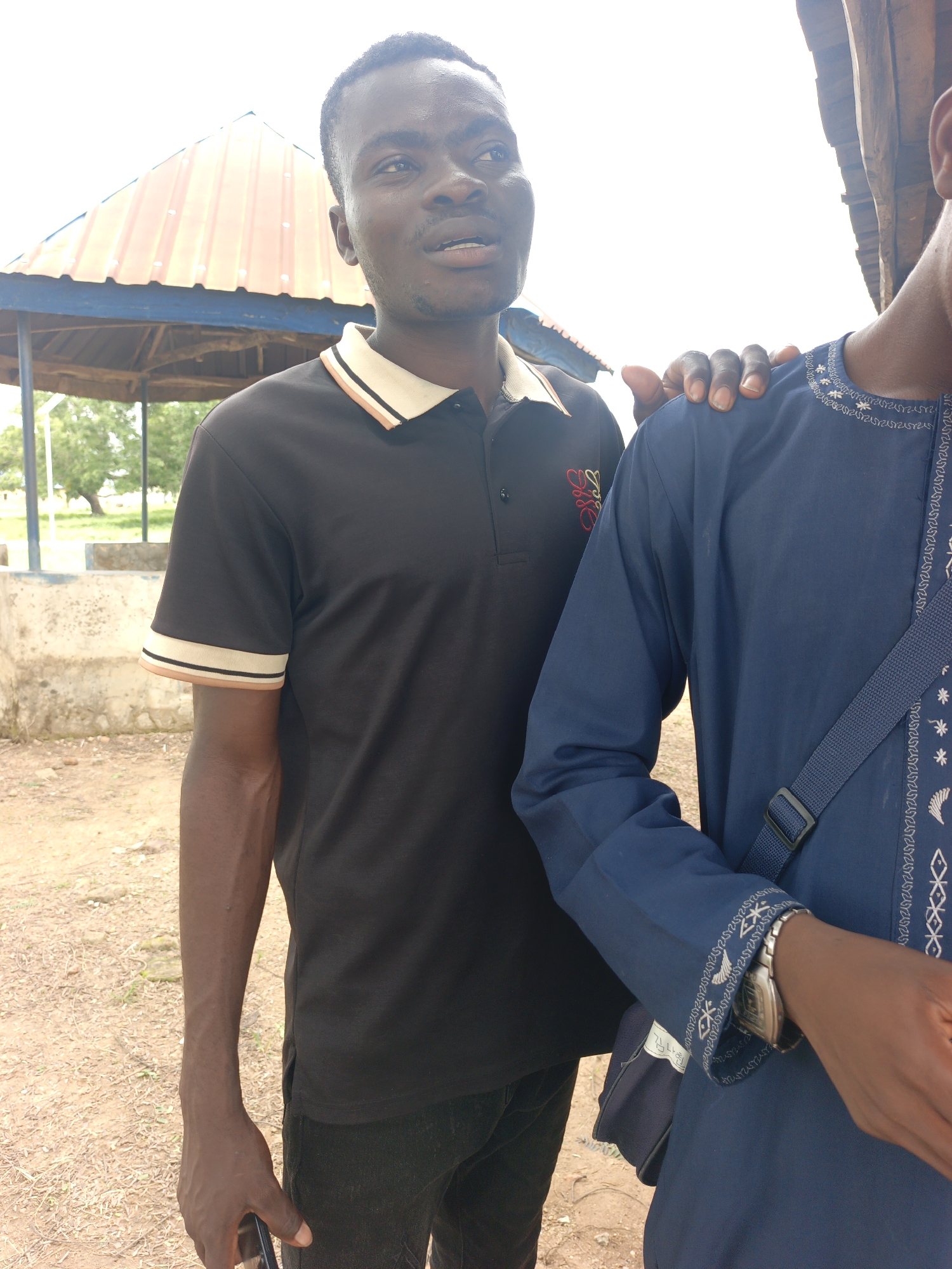 Two individuals are captured in a medium shot, standing outdoors. The person in the foreground on the left is a dark-skinned male wearing a black polo shirt with a tan and white striped collar and sleeve trim. He is looking off to his right with an open mouth and appears to be speaking. His left arm is visible, resting at his side, and he is holding a dark-colored object, possibly a phone, in his hand. The second person, on the right, is mostly obscured, with only their shoulder and upper arm visible. They are wearing a blue garment with intricate white embroidery along the neckline and sleeve. Their right hand is resting on the shoulder of the first individual. A dark blue bag with a strap is slung across their chest. The background features a rustic setting with a partially visible open-sided shelter with a corrugated metal roof, and some concrete structures. The ground is covered in dirt and gravel. The sky is overcast. The image suggests a casual encounter between two people in an outdoor, possibly rural or semi-urban, environment.