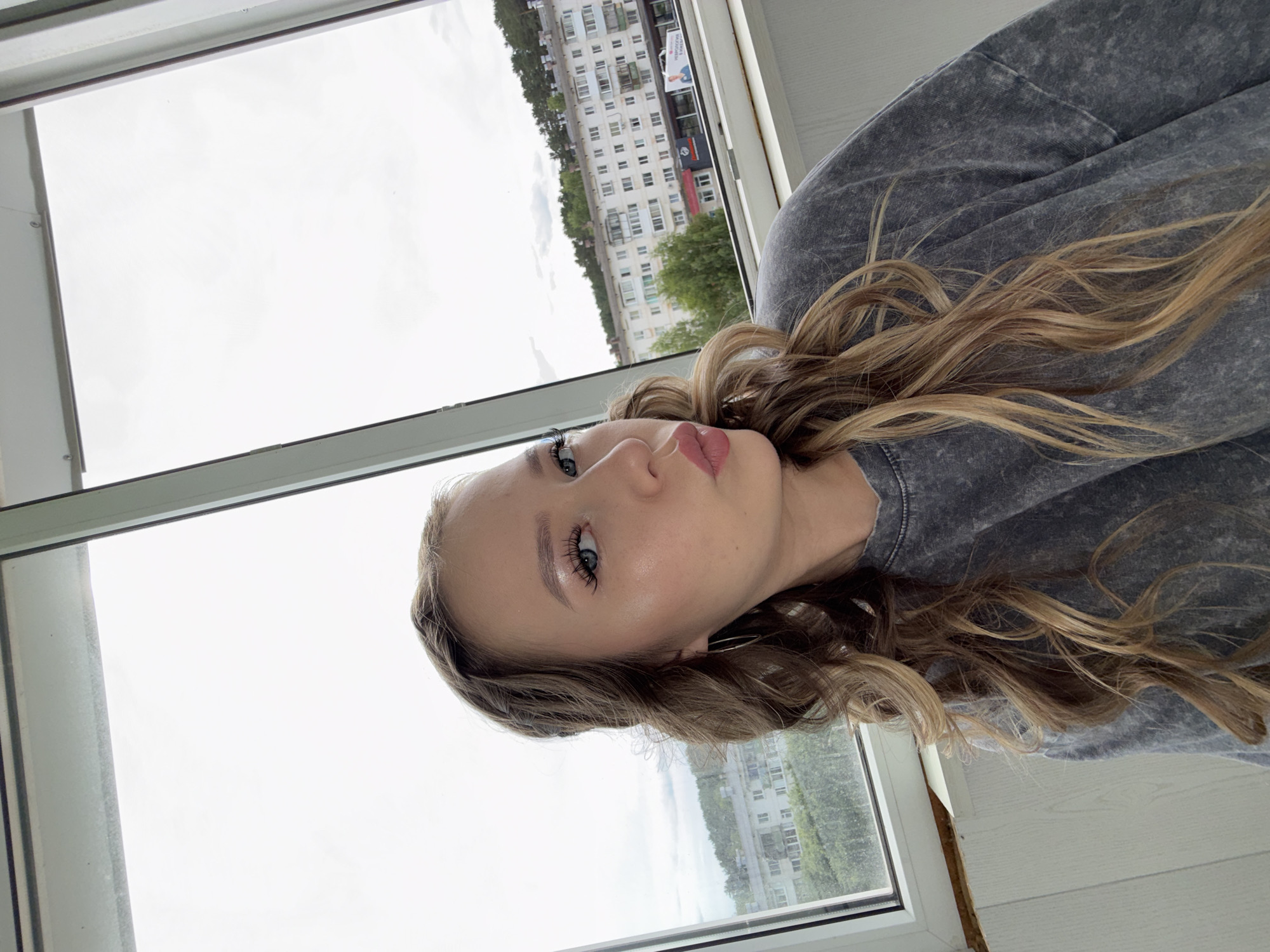 A young woman with long, wavy blonde hair and heavy makeup, including false eyelashes and lip gloss, poses for a selfie. She is wearing a grey, acid-washed crewneck sweatshirt. Her lips are pursed in a kiss pose, and she is looking at the camera. She has a silver hoop earring in her left ear. The background suggests she is indoors, near a large window that looks out onto a city street. In the distance, there are multi-story apartment buildings and a line of trees. The light in the image is overcast, consistent with a cloudy day. The visible cityscape aligns with the typical architecture and urban planning seen in Chaikovsky, Russia.