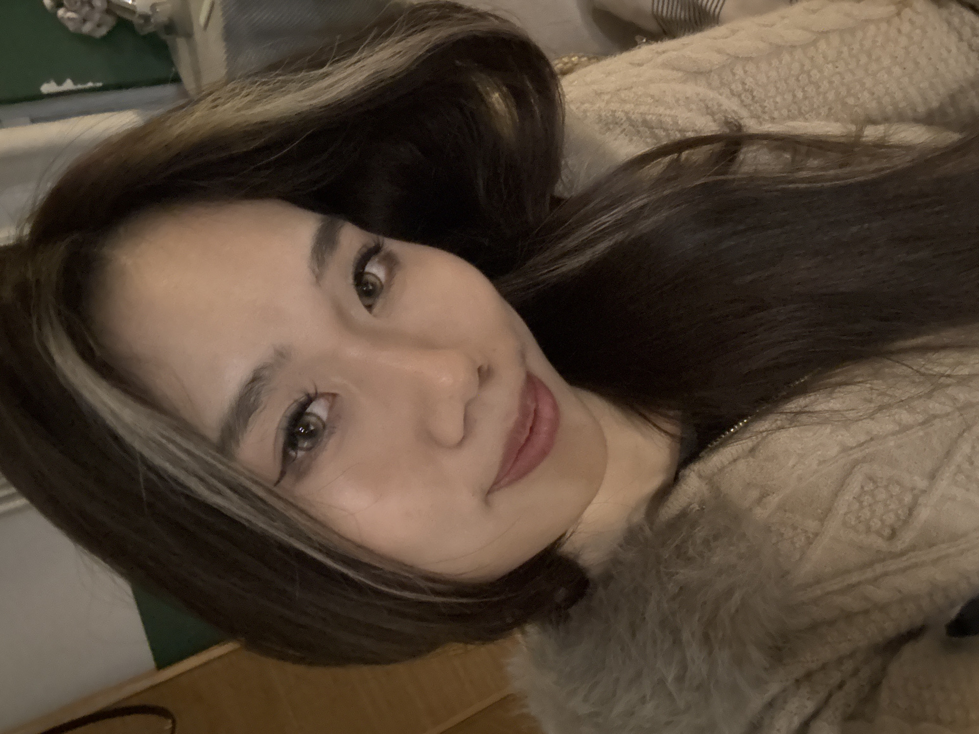 The image displays a close-up, chest-up portrait of an adult female taken indoors in the city of Liangjiang, China. The subject, appearing of East Asian descent, has long, dark brown hair with distinct light blonde or light brown highlights framing her face. She looks directly into the camera with a subtle, closed-mouth smile. Her eyes are light brown/hazel, accentuated by winged eyeliner and mascara. She is dressed in a beige or light brown cable-knit cardigan or jacket with a prominent, soft, faux fur-like collar of a matching color. A dark zipper is visible on the garment's front. The background is out of focus, revealing a vertical wooden panel and a dark green wall on the left, and a cream-colored patterned textile, possibly a blanket, and a blurry decorative object on the right. The lighting appears soft and artificial.