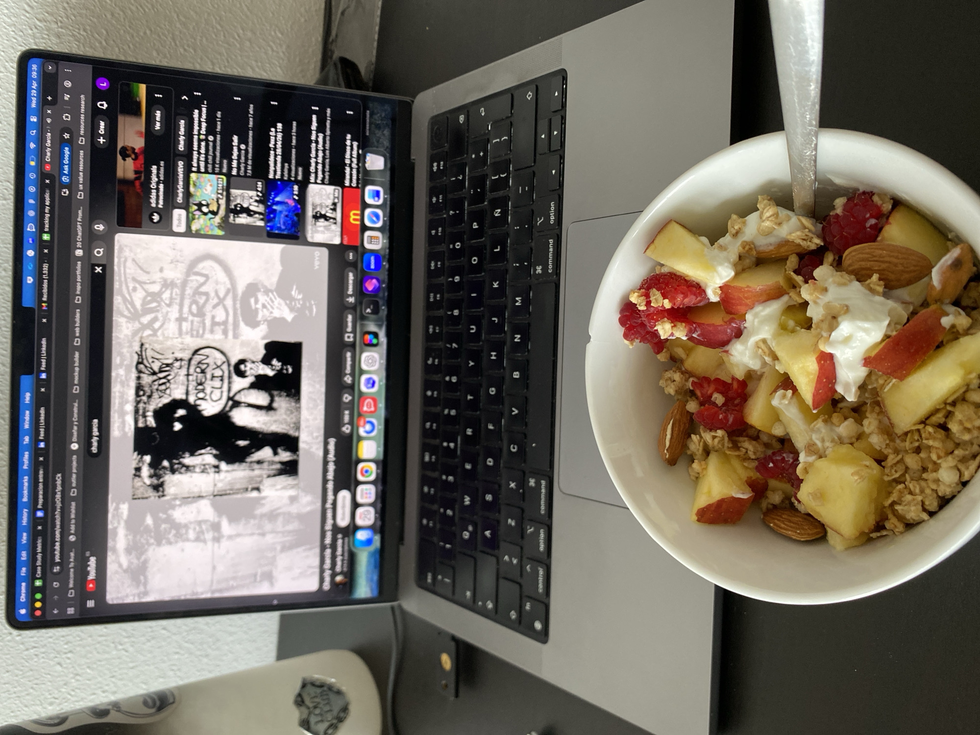 The image captures a morning scene, dated "Wed 29 Apr 09:36" on the screen, likely at an indoor location such as a home office or study area in Gijón/xixón, Spain. A silver laptop, appearing to be a MacBook Pro, is open on a dark grey or black desk surface.
Beside the laptop, a white bowl holds a breakfast or snack consisting of granola, yogurt, chopped red and yellow apples, raspberries, and whole almonds. A spoon rests within the bowl.
The laptop screen displays a web browser, likely Chrome, with numerous tabs open, including "YouTube," "LinkedIn," "Ask Google," "tracking my applicat...," "20 ChatGPT Prom...," "ux value resources," and "resources research." The primary content visible on the YouTube page is related to music, with "charly garcia" typed in the search bar. Featured videos include one with album art showing "MODERN CLIX TOYS" and titles such as "Charly Garcia - Nos Siguen Pagando Abajo (Audio)," "adidas Originals," "No Me Dejan Salir," and "Nonpalidece - Pexz." The YouTube interface also shows options like "Ver más," "Todos," "Descargar," and "Compartir."
The laptop's keyboard is a standard QWERTY layout, including Spanish characters like 'Ñ'. A portion of a white object, possibly a guitar or another device, with the text "LOVE CLUB" is visible in the bottom left corner of the frame. The scene suggests someone is having a meal while engaged in online browsing or work.