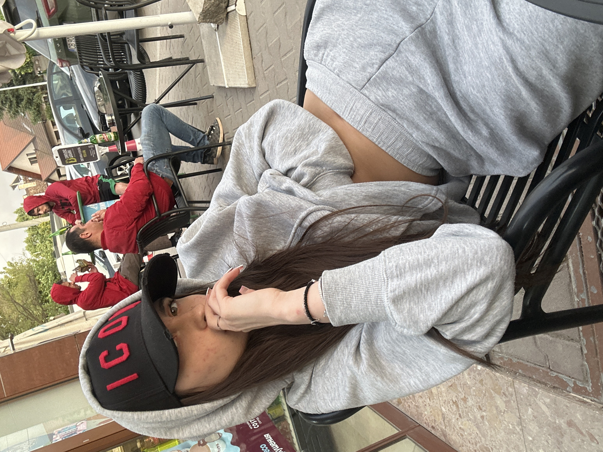 The image shows a young woman in a grey hoodie and "ICON" cap seated outdoors at what appears to be a cafe in Plovdiv, Bulgaria. The background features other people at tables, including two men in red jackets and another man in jeans. Drinks or bottles are visible on a table. A car is partially visible parked near the tables. The scene appears to be a casual, daytime outdoor setting.