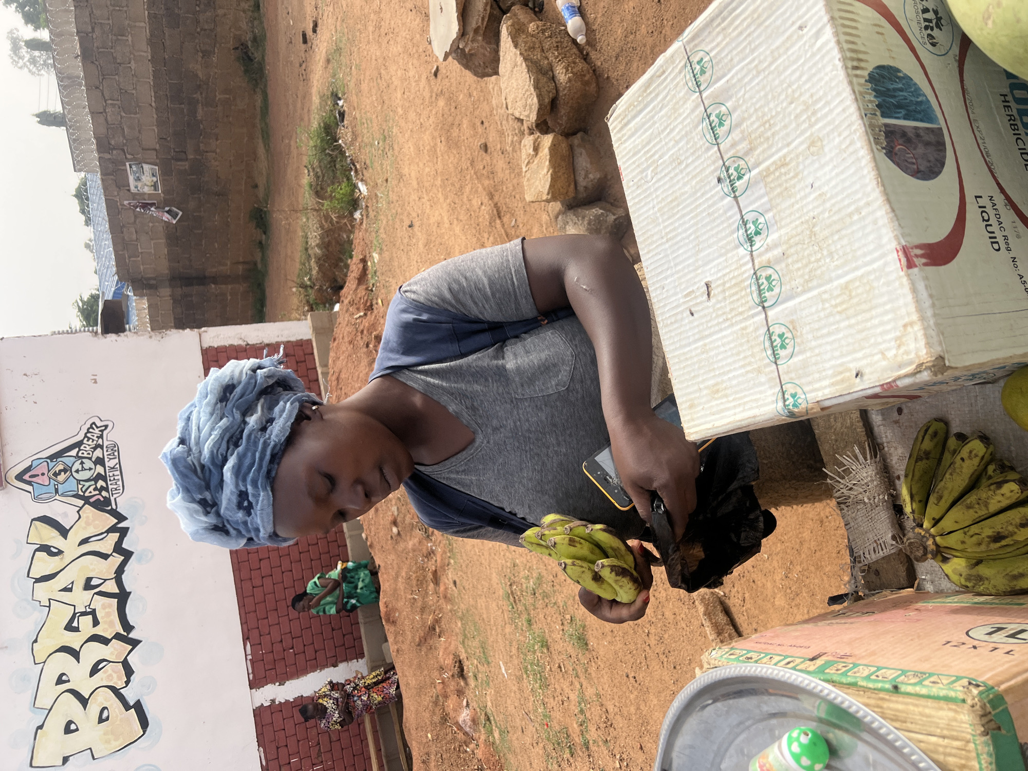 A woman wearing a blue headscarf and a gray t-shirt is preparing bananas at an outdoor market in Kirana, Nigeria. She holds a bunch of green and yellow bananas in one hand and appears to be cutting or peeling them with a small blade, possibly a pocket knife, held in the other. The woman is standing behind a display of cardboard boxes used as makeshift tables or counters. One box has the text "HERBICIDE LIQUID" and "SARO BIOSCIENCES" printed on its sides. Other bananas are on the boxes. In the background, there's a building with graffiti, a brick wall, and two other women sitting on the ground. The ground is reddish-brown dirt.