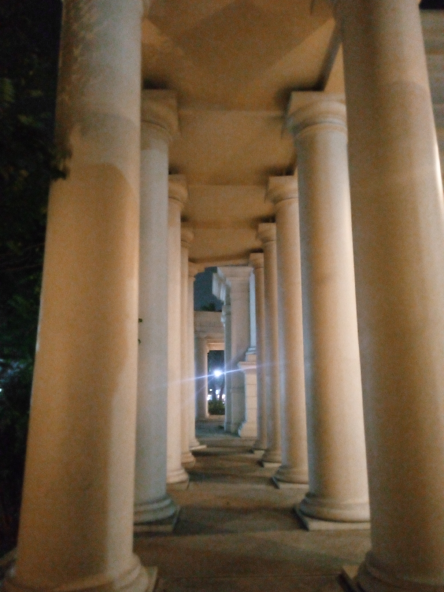 The image shows a perspective view of a colonnade at night. A series of tall, fluted columns line a pathway, receding into the distance. The columns are made of a light-colored material, possibly stone or concrete, and are illuminated by artificial light, creating warm highlights and soft shadows. The ceiling of the colonnade is visible overhead, also made of a similar material. In the background, there are lights from a building and possibly streetlights, suggesting an urban environment. On the left side of the image, a dark area with foliage indicates the presence of trees or bushes. The ground of the pathway appears to be paved, with some darker areas and a faint texture. There are no people or visible actions captured in the scene. The overall impression is one of an architectural structure at dusk or night. The specific location in the city of Bitung Jaya, Indonesia, is not discernible from the visual information provided in the image itself.