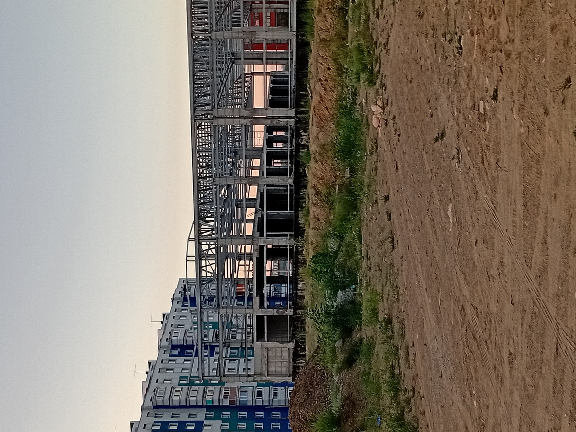 The image shows a construction site in the city of Semey, Kazakhstan, with a partially constructed building and a residential building in the background. The residential building, a multi-story structure with a white, blue, and green facade, is visible on the left side of the image. In the center and right of the image, a steel frame structure, characteristic of a new building under construction, is prominent. The ground in the foreground is dirt and sparsely covered with green vegetation. The sky above is clear and a light, pale blue, suggesting it is either early morning or late evening. There are no people visible in the image.