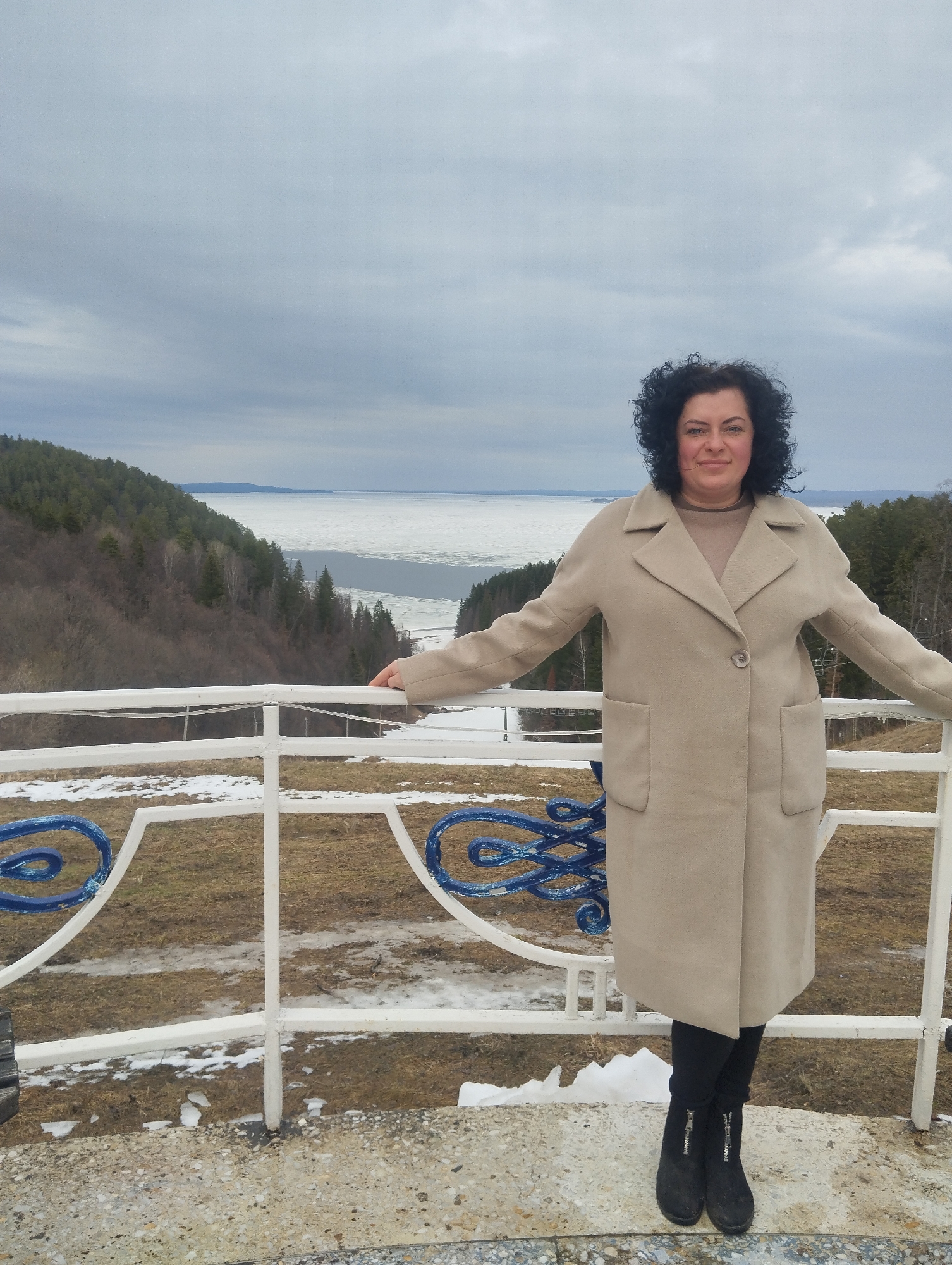 A middle-aged woman with dark curly hair stands in front of a white railing with blue decorative elements. She is wearing a tan coat, black pants, and black boots. She is resting her arms on the railing. Behind the railing is a landscape of trees and a body of water with ice. The location is Galevo, Russia. The sky is cloudy. There is snow on the ground.