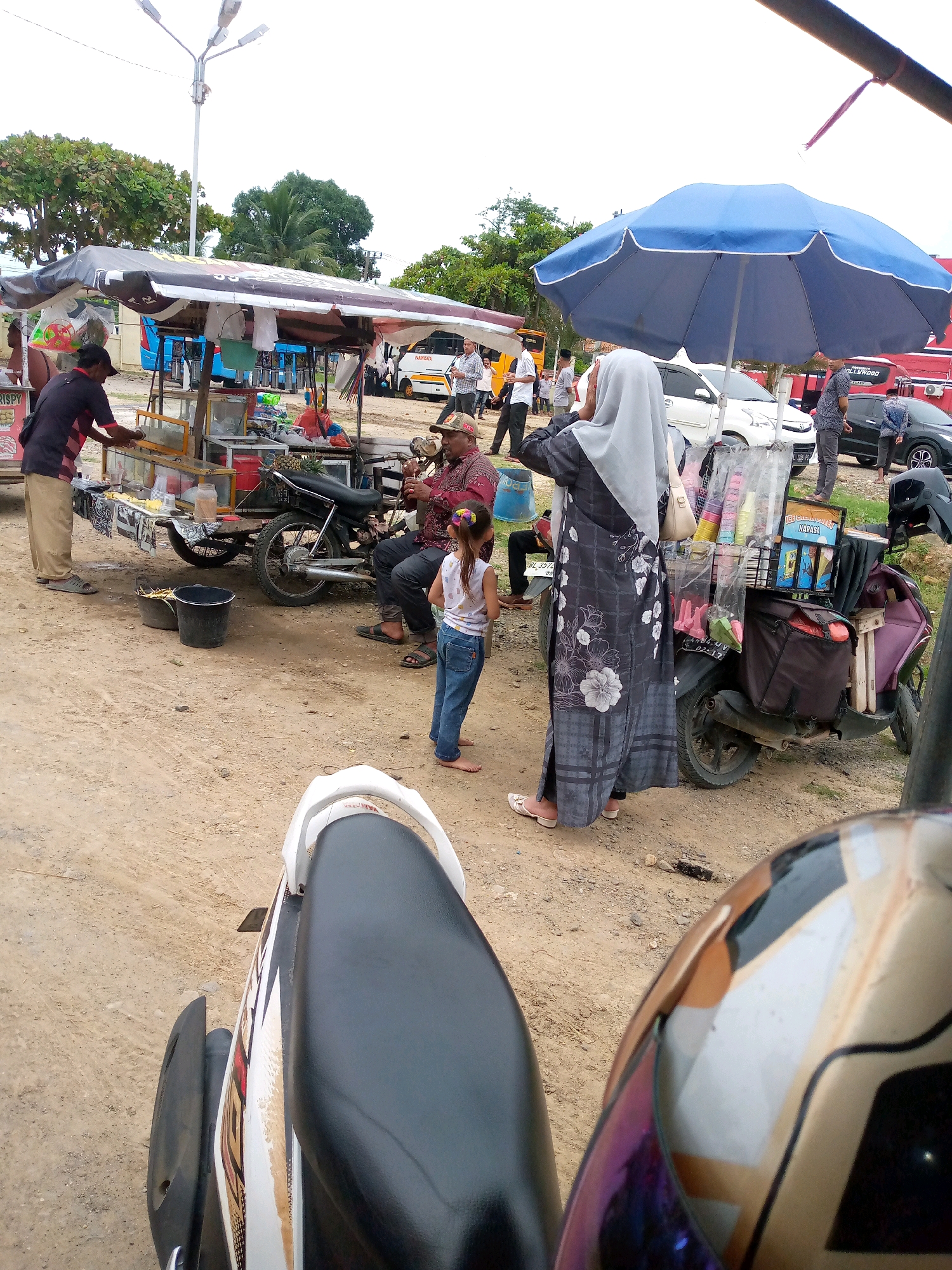 In Keudeteupinpunti, Indonesia, a street scene unfolds with several individuals and a mobile vendor. In the foreground, a motorcycle with a partially visible seat and helmet suggests the perspective of someone on a similar vehicle. The environment is a dusty, unpaved area.
To the left, a vendor stands behind a food cart, seemingly preparing or serving items. The cart is laden with various goods, including what appears to be fresh produce or prepared snacks. Adjacent to the cart, two black buckets are placed on the ground.
In the center of the scene, a man in a reddish patterned shirt, wearing a hat, is seated on a motorcycle and playing a stringed instrument, possibly a guitar or ukulele. A young girl in a white top and blue jeans stands near him, facing away from the viewer.
To the right, a woman wearing a grey hijab and a long, patterned grey dress stands near a blue umbrella. She is looking towards the food cart. Behind her, a motorcycle is parked, equipped with a rack holding an assortment of colorful items, likely drinks or packaged goods.
Further in the background, several vehicles are visible, including a white car, a bus, and a black car. More people are scattered around the area, suggesting a busy marketplace or gathering point. Trees and utility poles dot the landscape, indicating an outdoor urban setting. The overall atmosphere is one of casual commerce and community interaction.
