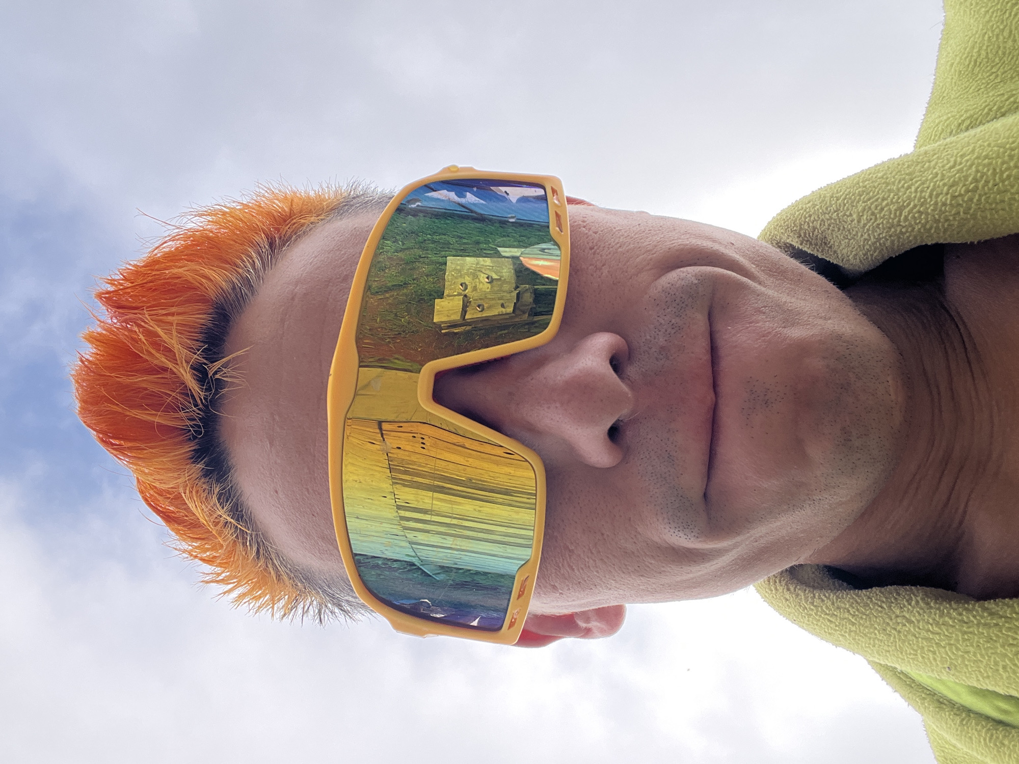 A close-up, low-angle selfie is taken of a person with bright orange, spiked hair and yellow wraparound sunglasses. The person is wearing a light green towel around their shoulders. The sunglasses reflect a wooden structure with vertical planks and possibly some greenery. The background consists of a cloudy sky. The image does not contain any elements that allow for the determination of a specific location within the city of Chaikovsky, Russia.