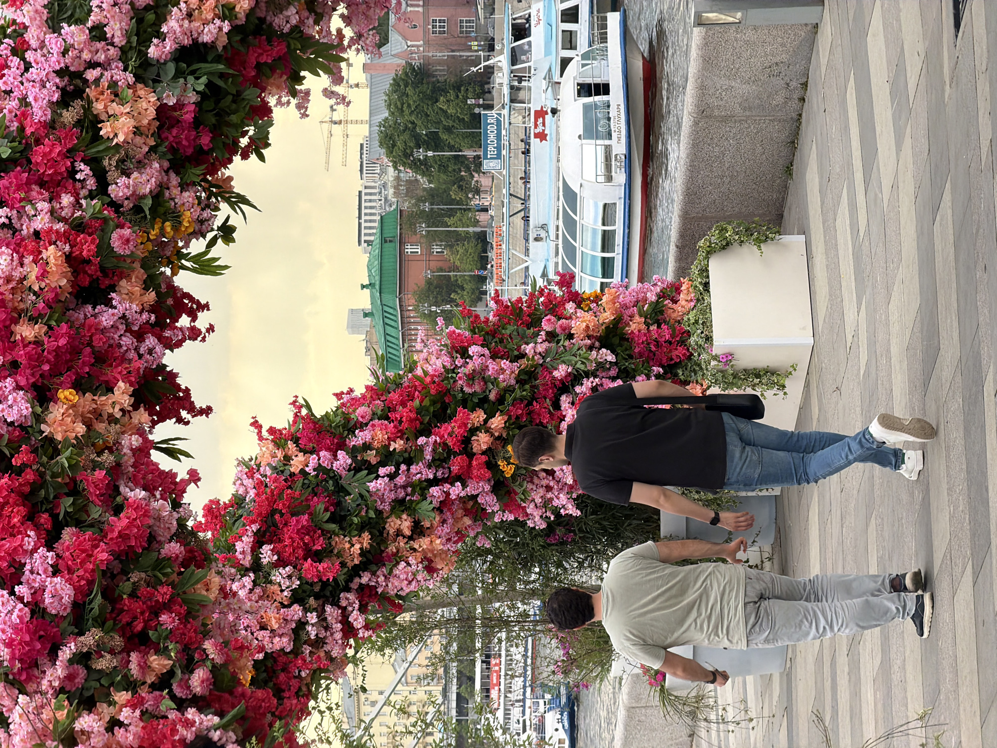 The image shows a flower arrangement on a city street in Unknown. The arrangement is pink, red, and orange, and is partially framing two people. The person in front wears a black shirt and blue jeans, while the person behind wears a light green shirt and light grey pants. The scene is set near a river or canal, as boats can be seen in the background. The location is identifiable as Russia due to the Cyrillic text on the boats. Buildings and a crane can also be seen in the background. The day appears to be overcast.