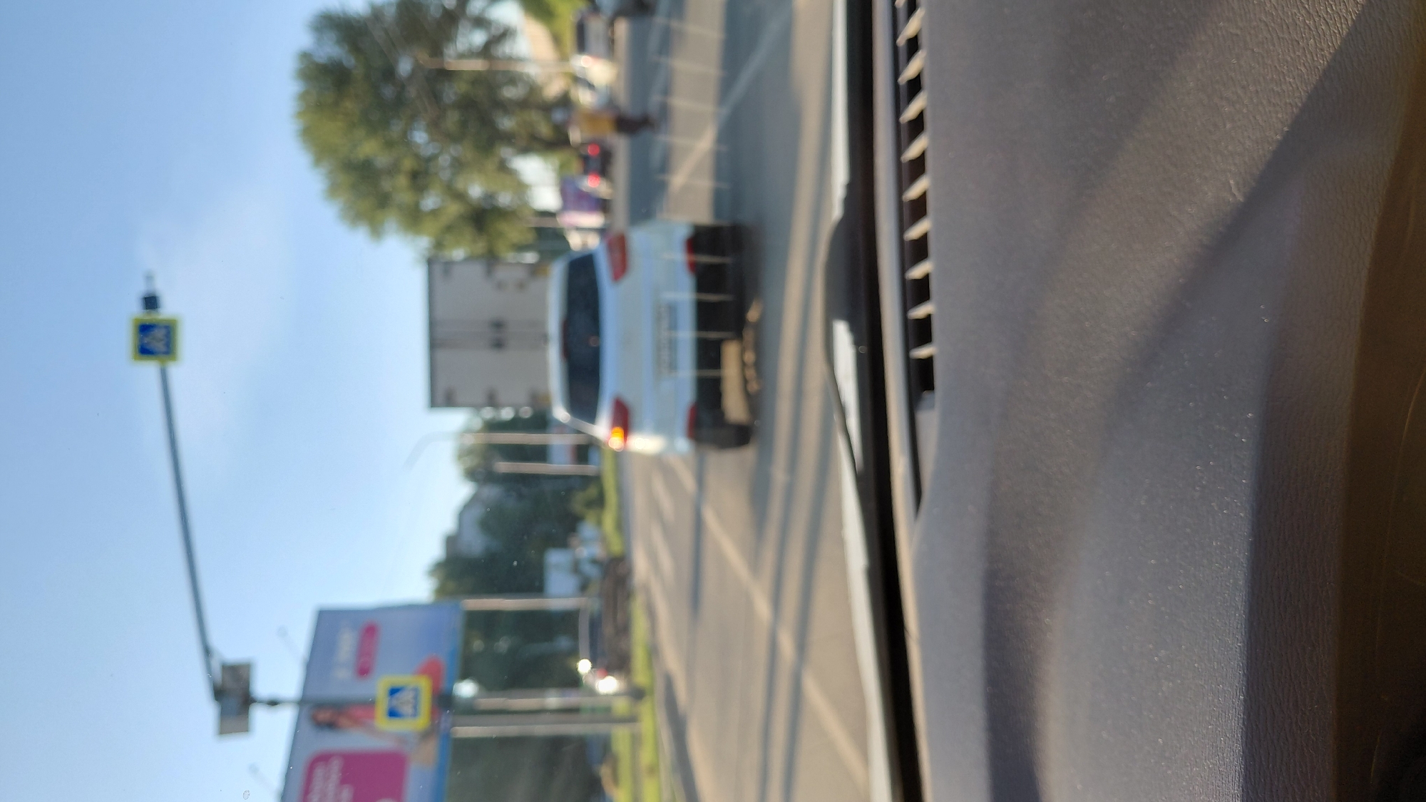 A view from inside a vehicle in Saratov, Russia, shows a white crossover SUV stopped at a pedestrian crossing. The SUV's brake lights are illuminated. A person wearing a yellow shirt is walking across the pedestrian crossing in the distance. In the background, a large truck with a white trailer is visible. Traffic lights and pedestrian crossing signs are mounted on poles. The scene is set against a clear blue sky and features trees and buildings. The foreground of the image is dominated by the interior of the vehicle, likely the dashboard.