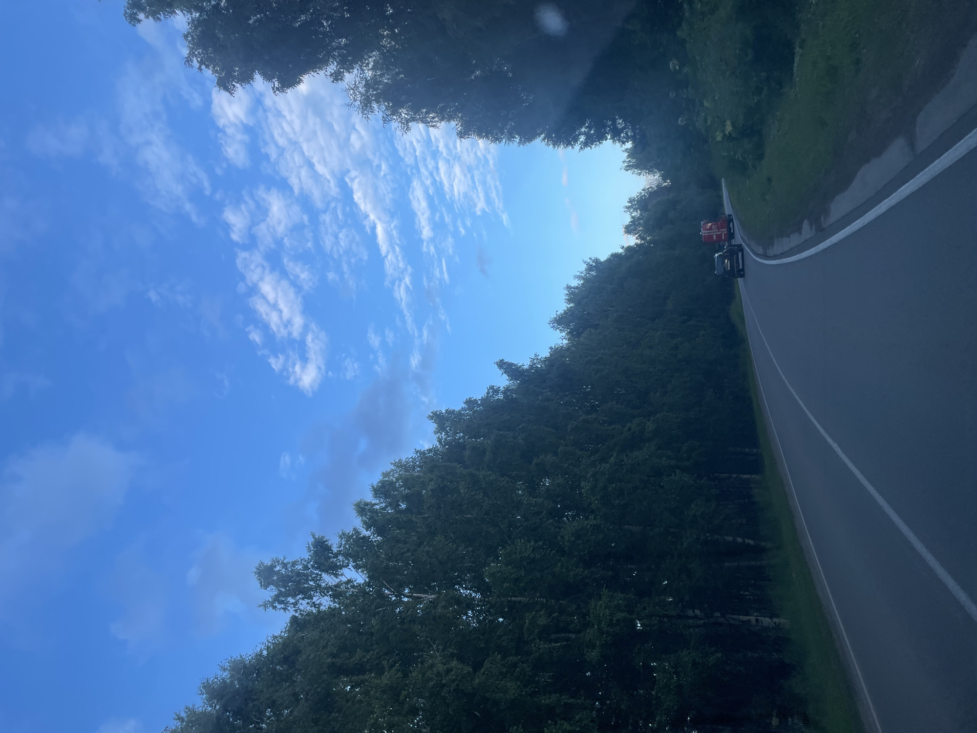 The image captures a view from a vehicle looking upwards and to the side, with the camera oriented vertically. The sky is a vibrant blue with scattered white clouds. Dense, dark green trees line the right side of the frame, forming a wall of foliage that obscures the ground behind them. A paved road curves gently into the distance on the right side of the image, bordered by a grassy shoulder and more trees.
Two vehicles are visible on the road. The first is a red truck, and behind it, a dark-colored vehicle, possibly a car or SUV. The scene suggests a travel scenario, possibly on a scenic route or a less-trafficked road given the surrounding nature.
The lighting indicates it is daytime, likely late afternoon or early evening, given the slightly muted sunlight and the deep shadows cast by the trees. The clouds suggest fair weather. The overall mood is serene and peaceful, with a sense of natural beauty and movement.
There is no visible text in the image. The location context, Камское, Russia, implies a rural or semi-rural setting within Russia.