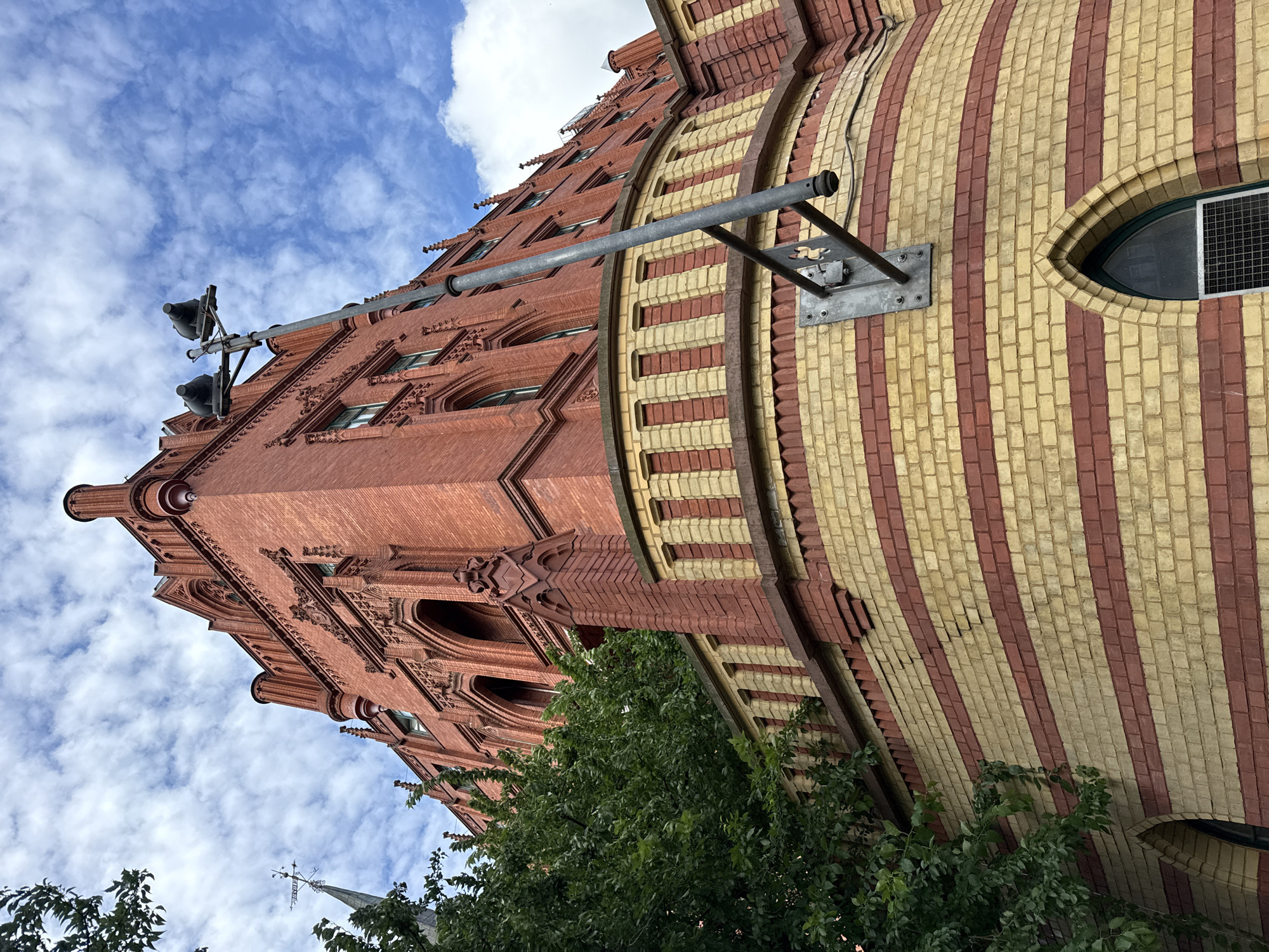An exterior, low-angle, full shot captures a red brick building with yellow accents, under a cloudy blue sky. The building's architecture features gothic-inspired arched windows and decorative brickwork along the roofline and facade. A prominent feature is a curved section of the building clad in horizontal bands of red and yellow bricks. A metal pole with two speakers or lights attached to the top extends from the building upwards into the sky. Lush green foliage from a tree is visible on the left side of the frame, partially obscuring the building. The building is likely located in Szczecin, Poland, given the architectural style and historical context often associated with the city. No people are visible in the image.