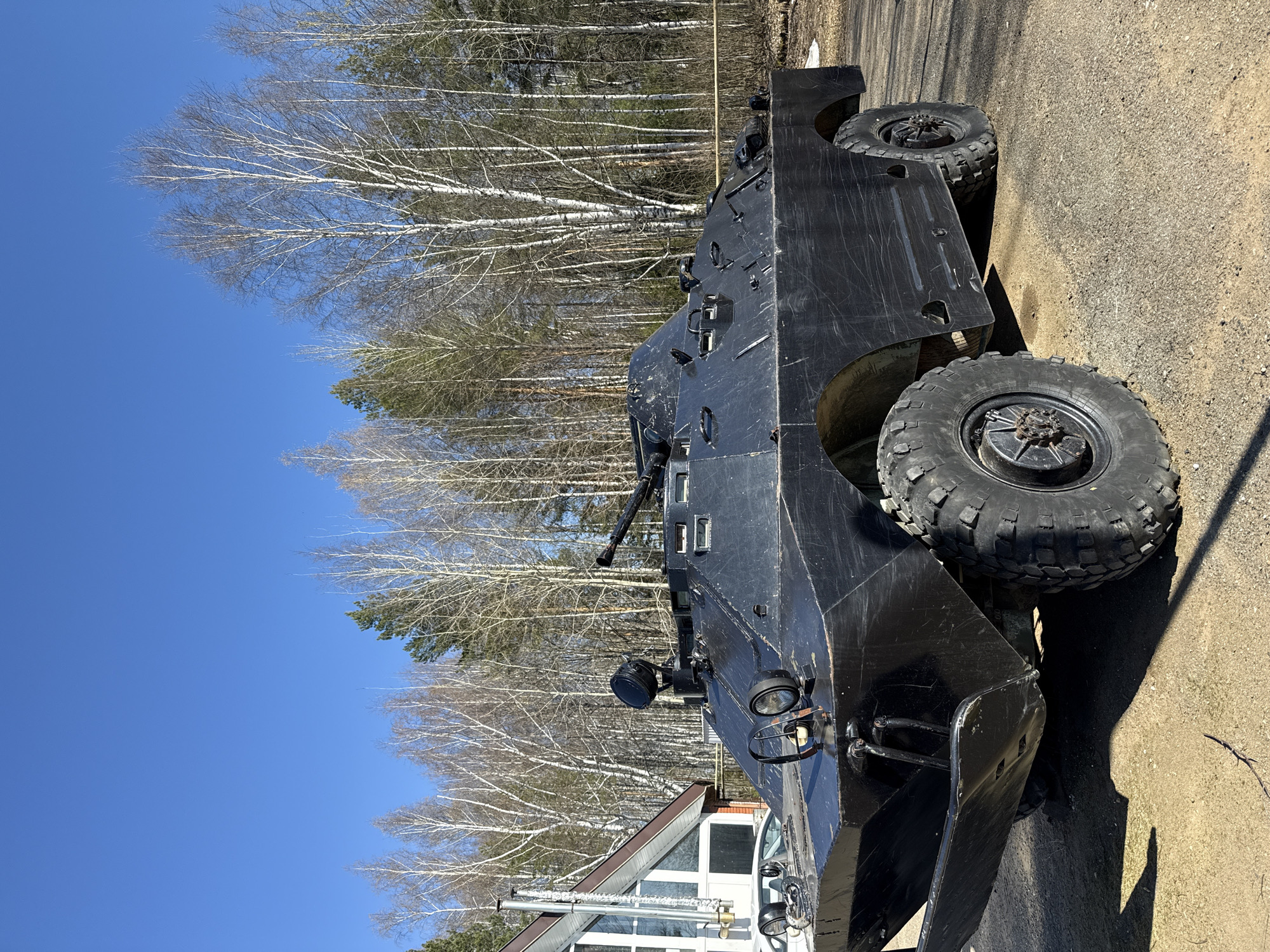 A black, wheeled armored vehicle is parked in an outdoor area, likely in the city of Галево, Russia. The vehicle has large, treaded tires and is situated on a dirt or gravel surface next to a paved area. A building with white-framed windows and a sloping roof is visible in the foreground, partially obscured by the vehicle. In the background, there is a stand of bare trees against a clear blue sky. The scene appears to be taken during daylight hours.