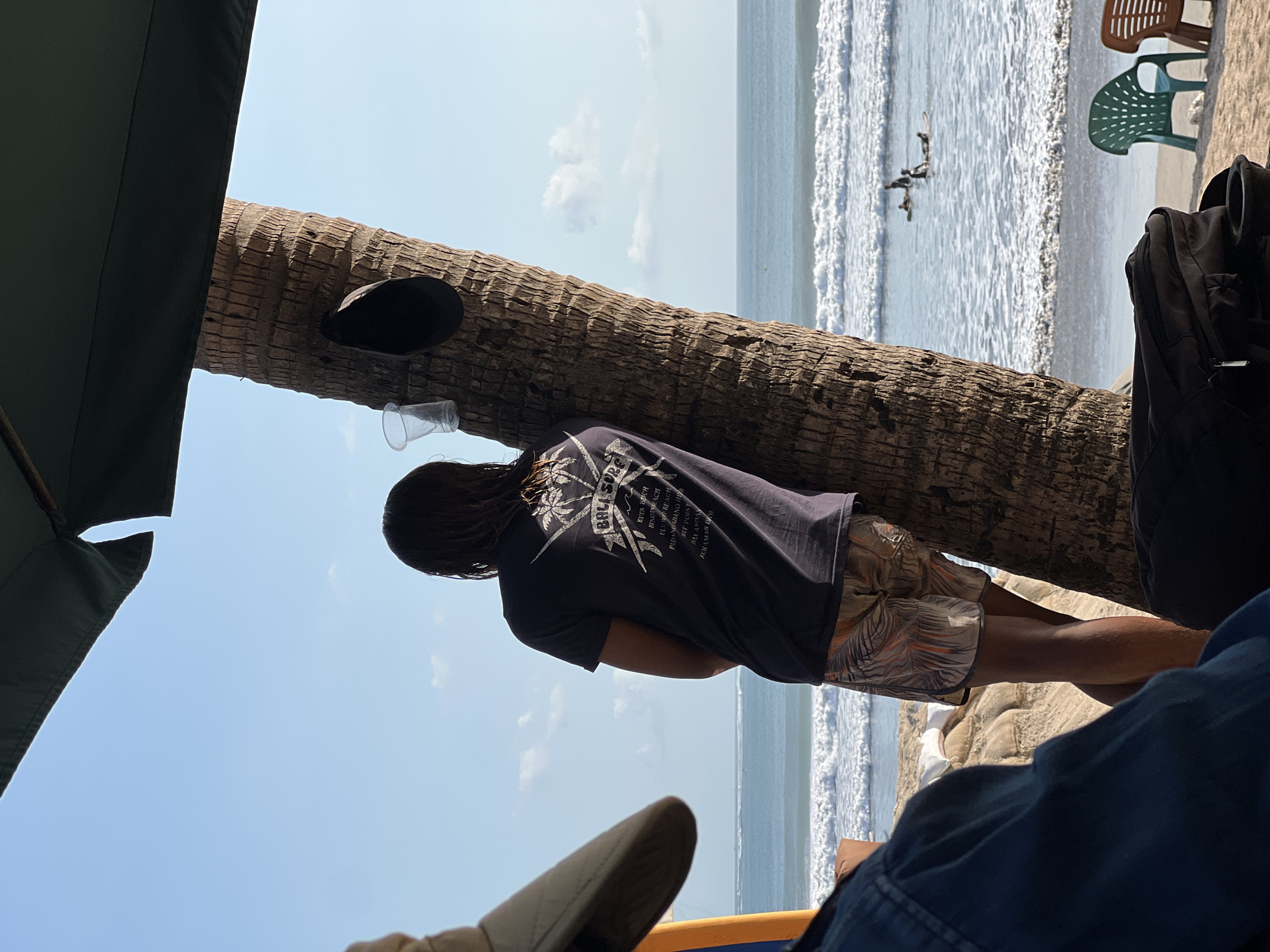 A man with long, dark hair, wearing a dark t-shirt with a "Bali Surf" graphic and patterned shorts, leans against a palm tree on a sandy beach. The ocean with breaking waves is visible in the background, with several surfers in the water. The scene is viewed from under a large, dark green umbrella, with parts of other beachgoers' attire and belongings in the foreground, including a blue denim jacket, a sun hat, and bags. Beach chairs are scattered on the sand. The image captures a scene in Kuta, Indonesia, a popular surf destination.