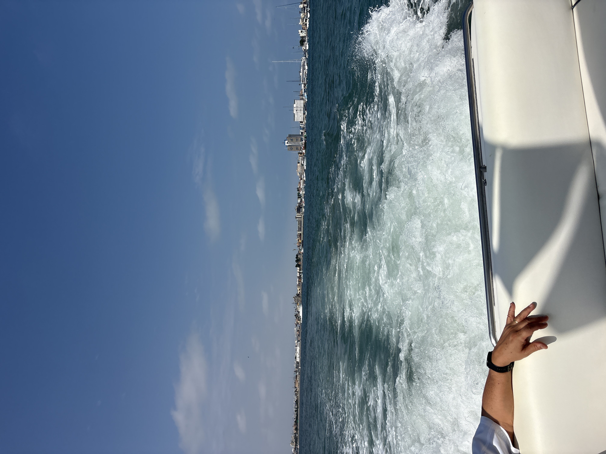 The image depicts a view from the stern of a moving boat, looking towards a coastline under a clear blue sky with scattered clouds. The boat's wake churns the turquoise water. On the left side of the frame, a person's arm and hand are visible, resting on the white upholstery of the boat. The person is wearing a white shirt and a black watch on their wrist, and a ring on one finger. In the distance, the coastline of Callao, Peru, is lined with buildings, including apartment complexes and other structures, and a marina filled with sailboats and yachts. The scene conveys a sense of movement and leisure on the water, with the urban landscape of Callao serving as a backdrop.