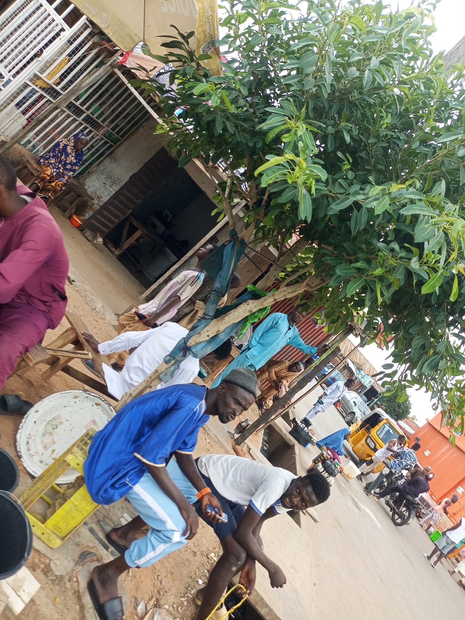 The image depicts a street scene in Arawa, Nigeria, with several individuals and objects. A woman in a blue and gold patterned dress is seated under a corrugated metal structure, which appears to be a shop or market stall. In the foreground, a man in a blue shirt and light blue pants is crouched down, looking at his phone. Another man in a white t-shirt is also visible in the foreground, his head turned towards the camera with a smile.
Various objects are scattered around the scene, including buckets, crates, and a large metal plate. A tree with lush green leaves shades part of the area. In the background, a yellow tricycle, a motorcycle, and several other people can be seen, suggesting a busy urban environment. The overall impression is one of a typical day in a Nigerian marketplace, with people engaged in daily activities.