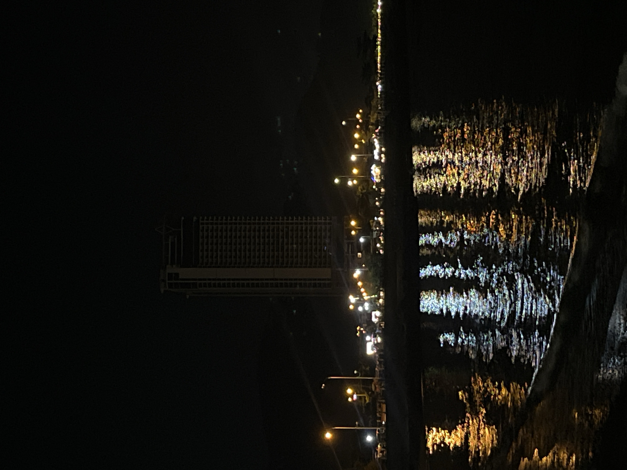 Captured at night, the scene features a body of water with the reflections of city lights shimmering on its surface. Along the waterfront, streetlights illuminate the path. In the background, a multi-story building stands tall, its facade dotted with lighted windows. This view is suggestive of a location within or near Bac Nha Trang Ward, Vietnam, known for its urban coastal environment.