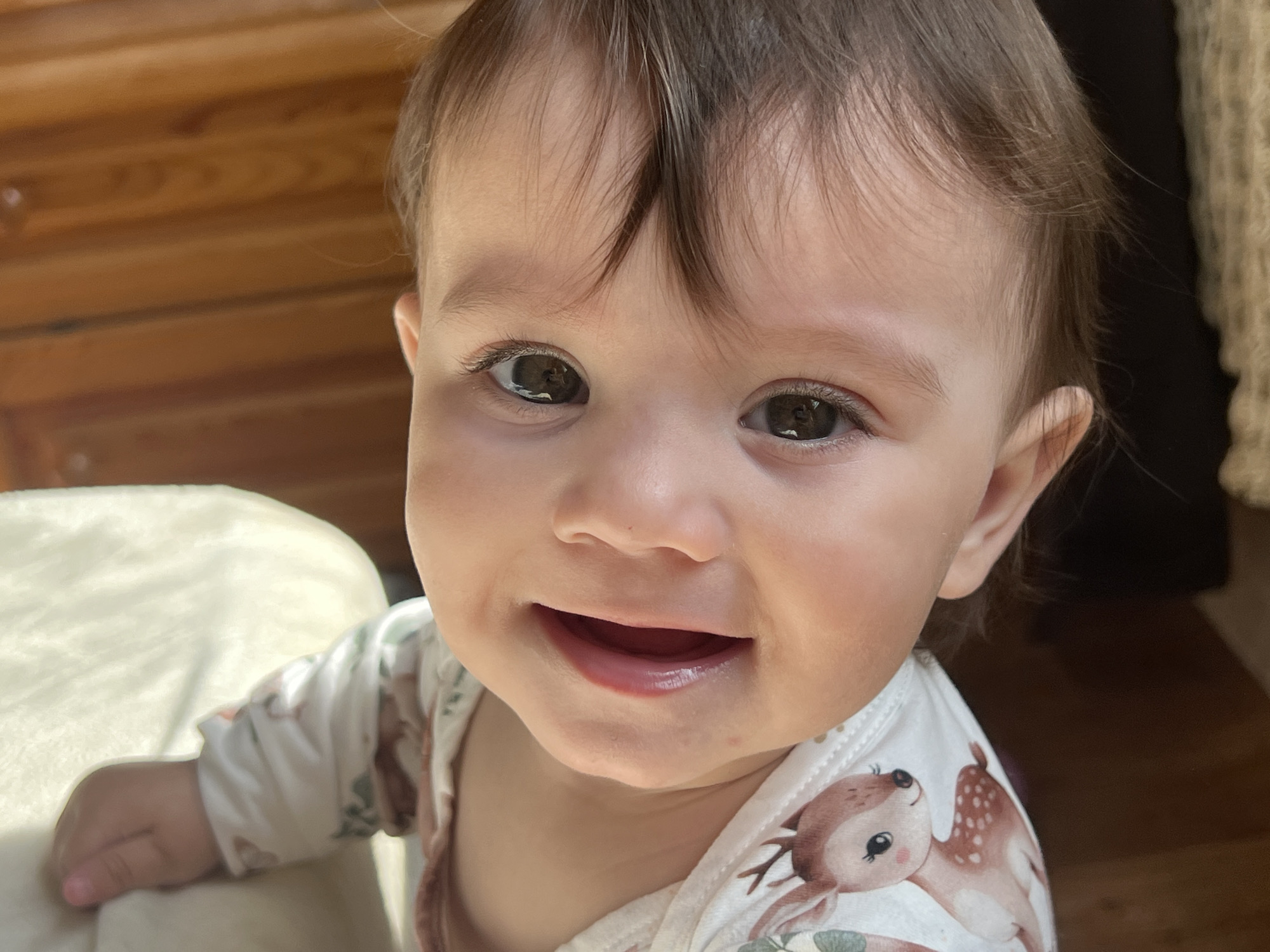 A close-up, frontal image depicts an infant subject. The infant has light skin, dark brown eyes, and short, brown hair with bangs. The mouth is open in a wide smile, with no visible teeth. The subject is wearing a white, long-sleeved garment featuring a repetitive pattern of light brown fawns and green botanical elements. To the left and upper background, blurred horizontal wooden planks are visible. The right background is dark, with a textured, light brown knitted or woven material partially visible. A light-colored, soft surface supports the infant's left arm in the lower-left foreground. The scene is brightly illuminated, indicating an indoor setting in the city of Elena, Bulgaria.