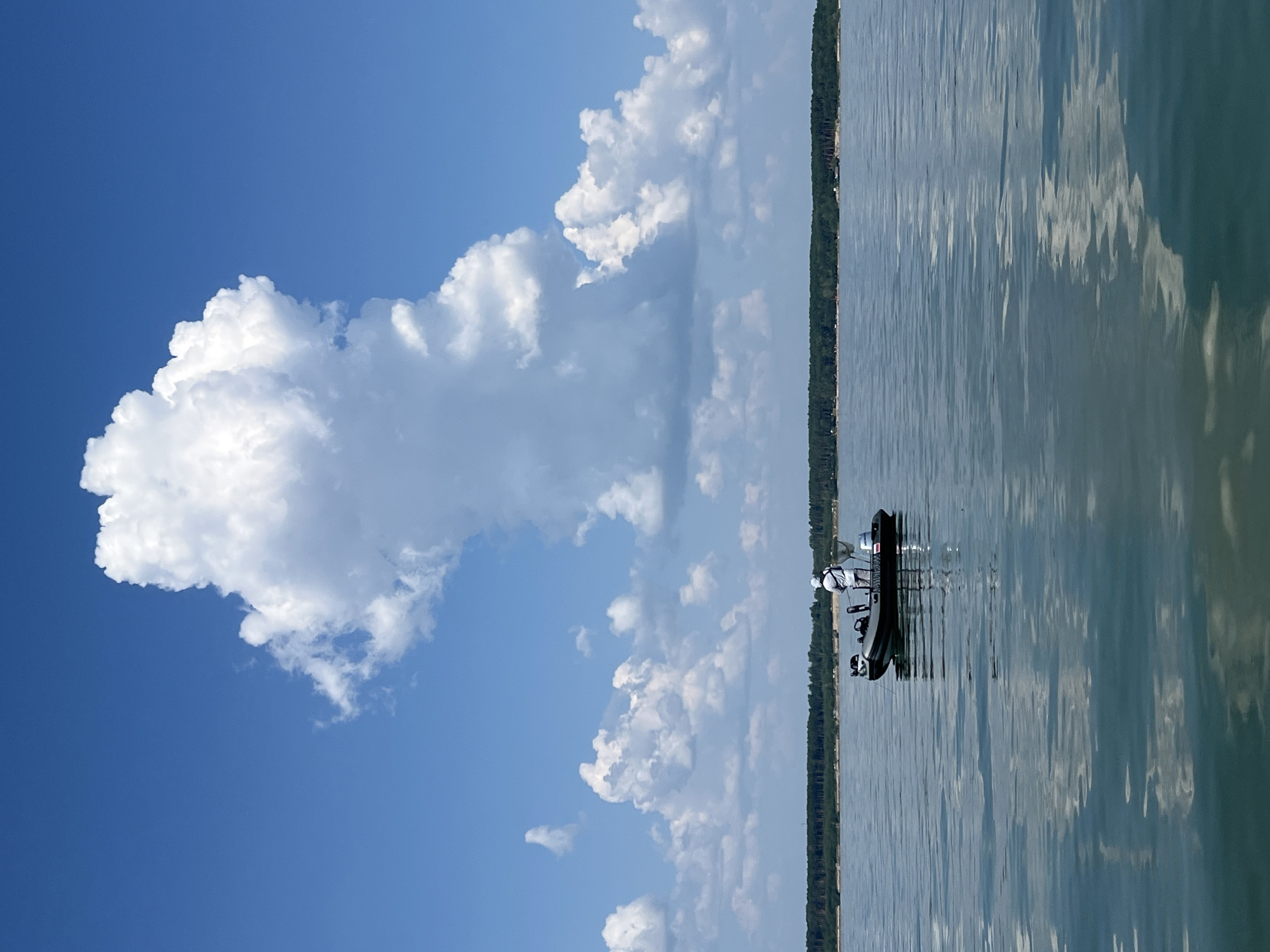 The image depicts a calm body of water with a distant treeline along the horizon. A small, dark-colored boat with two outboard motors is positioned in the middle of the frame. A single person, wearing a white hat and shirt, stands in the boat, appearing to be engaged in fishing. The sky is a vibrant blue, dotted with large, fluffy white clouds. The water reflects the sky and clouds, creating a shimmering effect. The overall scene suggests a peaceful, sunny day on a lake or river. The location is identified as Borovoe, Russia.