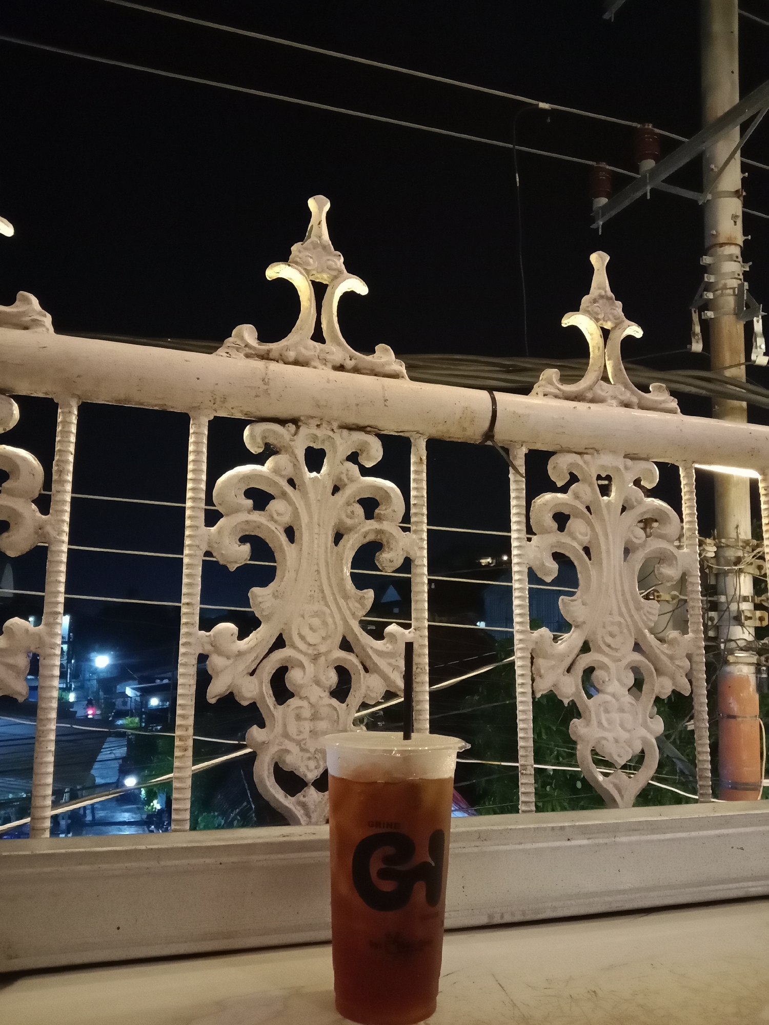 A tall plastic cup of iced tea with a black straw sits on a white ledge in the foreground. The cup has a black logo with the letters "GH" and the word "DRINK" visible. Behind the cup, a white decorative metal railing with intricate scrollwork separates the viewer from a nighttime city scene. In the background, streetlights and building lights illuminate the area, suggesting a commercial or residential district. Electrical wires and poles are visible above the railing, and the sky is dark. The overall impression is of a beverage enjoyed outdoors at night. The location is identified as Pandean, Indonesia.