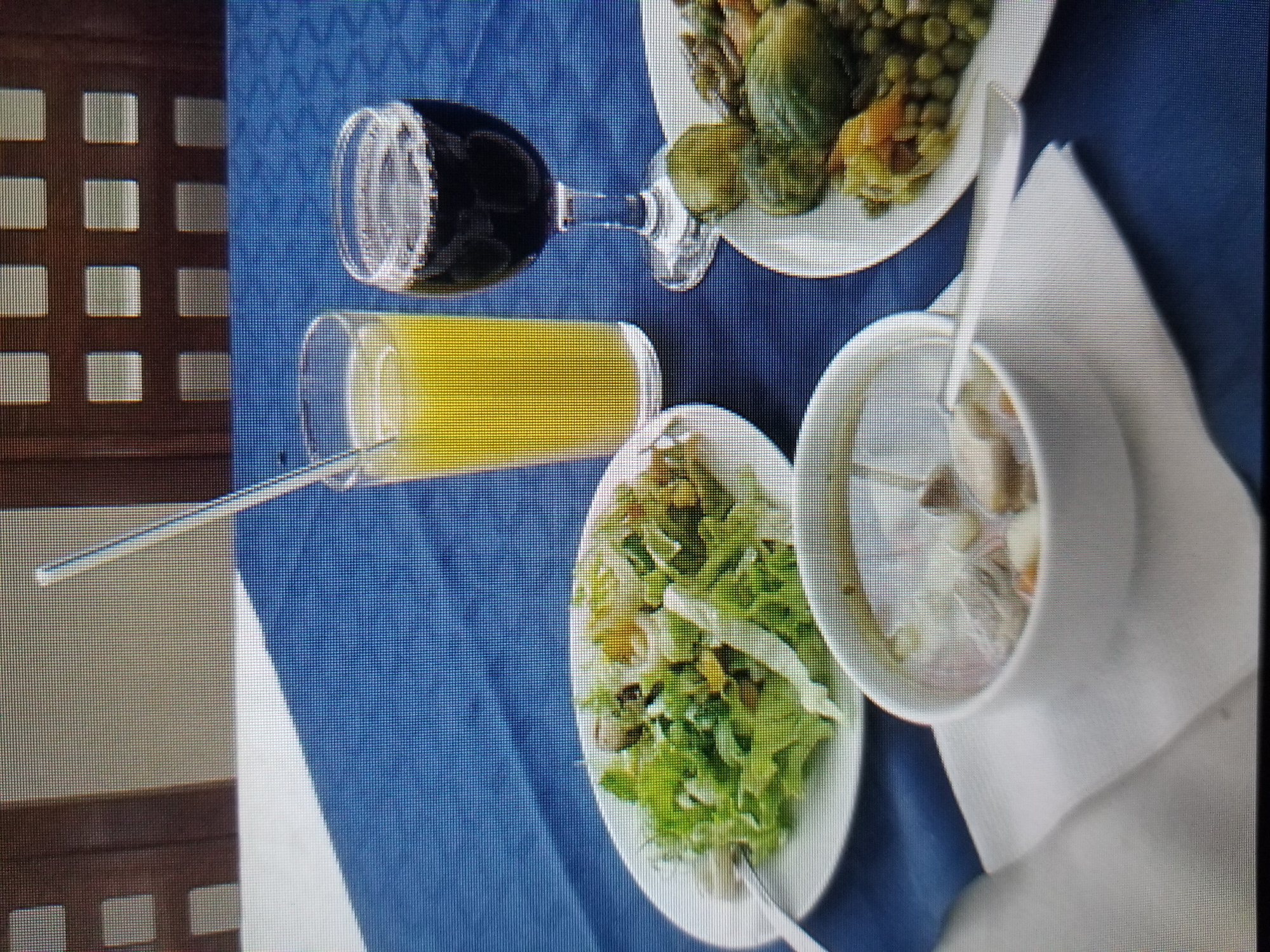 The image displays a table set for a meal on a blue tablecloth. On the table, there is a white plate with a green salad, a white bowl of soup, a glass of yellow juice with a straw, and a glass of dark red liquid. To the right of the red liquid, there is another white plate with a dish containing green vegetables and what appears to be small potatoes or peas. In the background, there are wooden chairs with a grid-like pattern. The scene appears to be outdoors or in a brightly lit indoor setting. There is no indication of the specific location in Tiraspol, Moldova, nor are there any people or actions depicted.