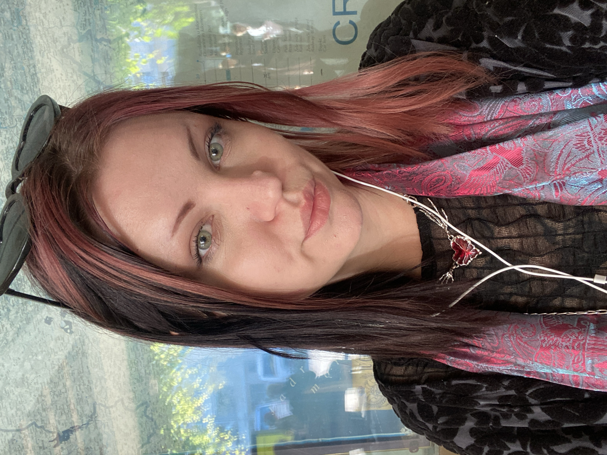 The image shows a woman with light skin, green eyes, and dark hair with red highlights. She is wearing a black top with a floral pattern and a pink and blue patterned scarf. She also has sunglasses on her head and white earphones around her neck. A silver chain with a red pendant is visible. The background appears to be a map or display, possibly inside a vehicle like a bus or tram, with text that includes "Razdaljina u kilometrima," "kilometers," and "CF" (likely standing for "Car Ferry"). The text is partially obscured but gives hints about the location being Montenegro, as the language suggests, possibly the city of Budva. The woman seems to be looking directly at the camera.