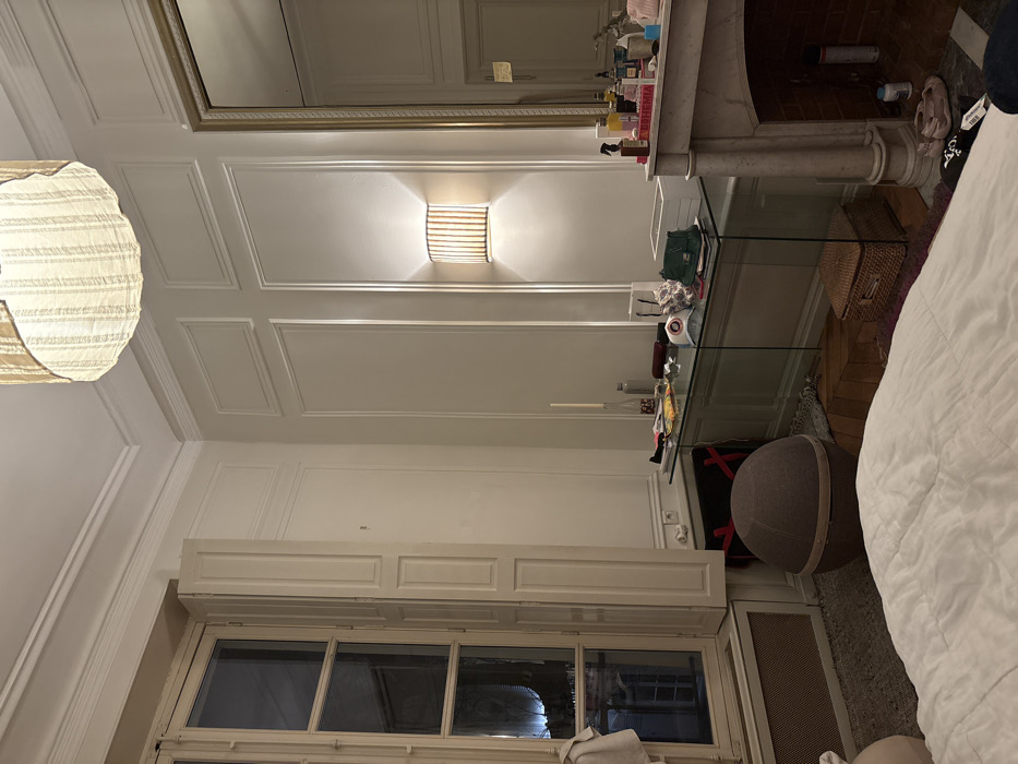 The image displays an interior room, indicative of a classic Haussmannian apartment in Paris, France, identified by its high ceilings, ornate crown molding, and decorative wall paneling. The scene is illuminated by dim, warm artificial light, suggesting evening or night.
In the foreground, the edge of a bed covered with a white duvet is visible. To the left, a tall, multi-paned window features internal bi-fold shutters, one half partially open, revealing a dark exterior with reflections of structures. A radiator is positioned beneath the window. A white towel or cloth hangs over the window frame.
Against the right wall, a glass-topped console table holds numerous personal items, including bottles, a tall white candle in a bottle, a white baseball cap with a logo, a green handbag, and a white paper bag. Above the console, a rectangular wall sconce with a striped shade emits light upwards and downwards. To the right of the console, a classical marble fireplace with a brick interior is present. Its mantelpiece is crowded with various bottles, sprays, and cosmetics, including a pink item and an object clearly labeled "BOHEMIA." A large, gold-framed mirror is mounted above the fireplace, reflecting portions of the room.
On the floor near the window, a large, grey spherical object with a central band, resembling an ottoman or exercise ball, is situated. A woven rectangular basket rests on the floor beneath the glass console table. A large, drum-shaped ceiling light fixture with a striped fabric shade hangs from the ceiling. The floor features a rug partially covering wood flooring. No individuals are present in the scene.