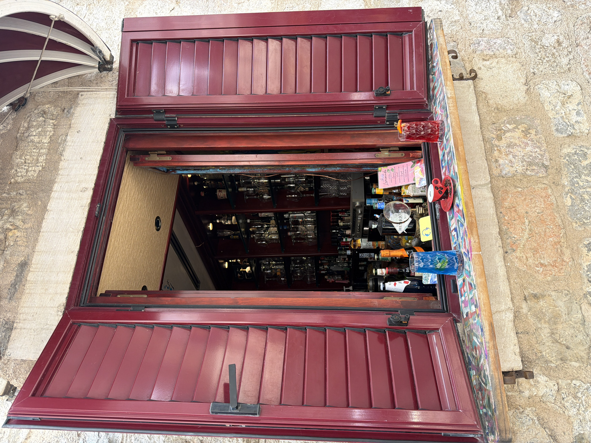 The image shows a view into a bar through an open window with red shutters. Inside the bar, shelves are lined with bottles of alcohol and glassware. On the windowsill, there is a blue glass with a drink and a small red cup with coffee beans. To the right, there is a red drink in a glass with an orange garnish. The bar appears to be located in the old town of Budva, Montenegro, given the stone building material and the style of the awning visible at the top right. The scene is set during the day.