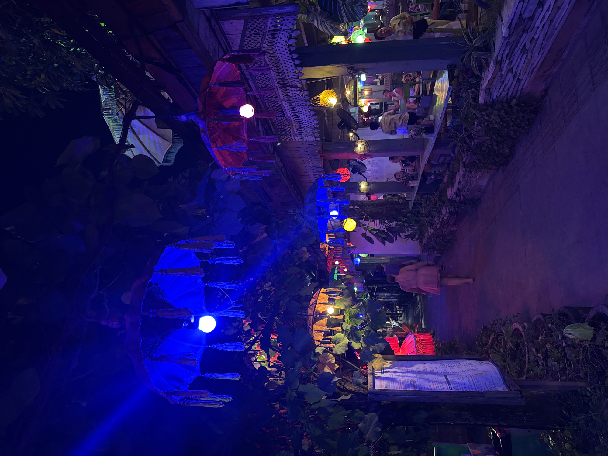 The image depicts a street scene at night, likely in Kuta, Indonesia, given the vibrant and tropical ambiance. The foreground features a pathway illuminated by the ambient glow of various colored lights, predominantly blue and purple, creating a mystical atmosphere. Lush green foliage drapes overhead, forming a natural canopy from which decorative elements are suspended. These include numerous ornate umbrellas, some in striking blue and red, adorned with tassels and lit from within by bright bulbs. Further along the path, a woman with her back to the camera, wearing a light-colored dress, walks away from the viewer. The pathway leads to a restaurant or bar area on the right side of the image. This area is characterized by wooden structures, balconies with intricate carvings, and more colorful lights, including yellow lanterns and globe lights. Several patrons are seated at tables, some engaged in conversation. The overall impression is of a lively and colorful nighttime entertainment district, possibly a popular tourist spot in Kuta.