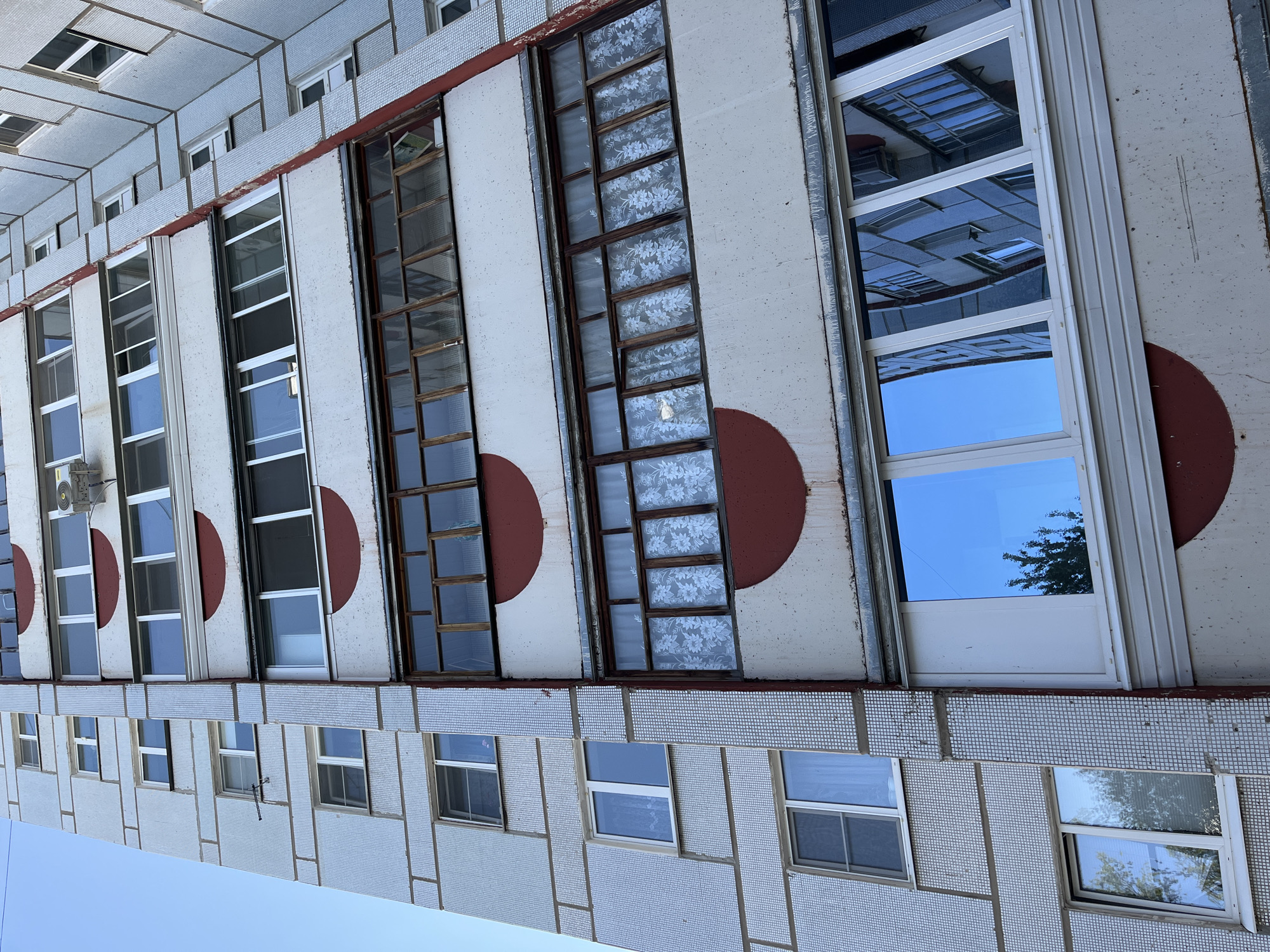 The image depicts the exterior of a multi-story building, likely an apartment complex, in Chaikovsky, Russia. The building's facade features a tiled lower portion with small windows and a higher section with larger, vertically-oriented windows. The larger windows are set within white frames and interspersed with prominent, semi-circular red designs. Some windows have clear glass, reflecting the sky or surrounding structures, while others have curtains or coverings. An air conditioning unit is visible mounted near one of the windows. The perspective is slightly angled, showing the building from below and tilted to the left. The sky is visible in the lower portion of the frame.