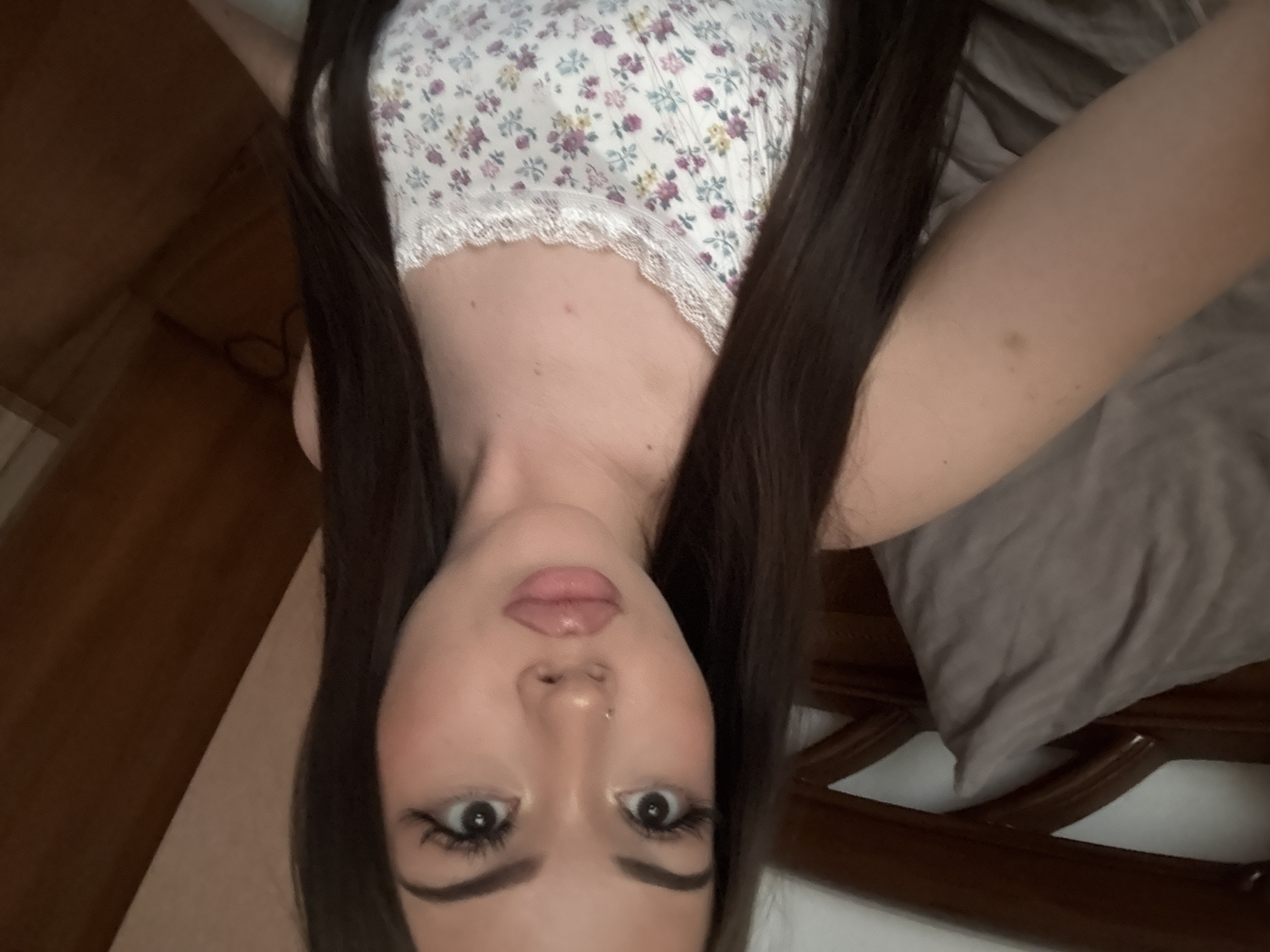 A close-up, inverted portrait of a young person, likely female, is captured indoors in Naberezhnye Chelny, Russia. The subject's face is central to the frame, viewed upside down, suggesting the photograph was taken while she was lying down. She has long, straight dark brown hair that frames her face, prominent dark eyebrows, large dark eyes with noticeable eyelashes, and full, light pink lips, accompanied by a subtle nose piercing. She is wearing a light-colored top adorned with a delicate floral pattern in shades of purple, yellow, and green, finished with white lace trim. Her gaze is direct and neutral towards the viewer. The background on the left side shows a dark brown wooden surface, possibly part of furniture, while the right side reveals a grey pillow or bedding and a dark brown wooden bed frame with a slatted design. The soft, even lighting indicates an indoor setting. The image depicts a direct portrait or selfie, with no specific activity beyond posing. There is no visible text within the image.