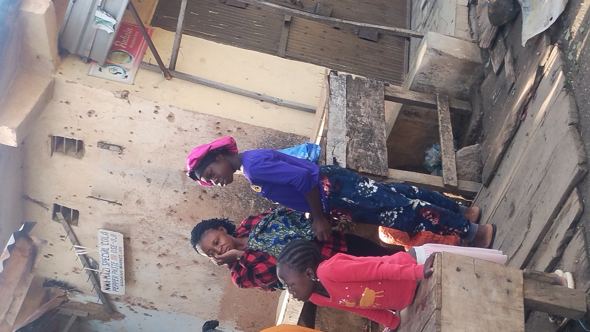 Three females of varying ages are positioned next to a weathered wooden structure. The oldest female wears a pink head covering, a purple top, and floral-patterned bottoms. The middle female wears a red and black plaid top. The youngest female is in a red long-sleeved top. In the background, a partially deteriorated wall displays a sign reading "NWA MAZI SPECIAL COLA PEPPER PASTE OR OSE-OJI GADA SU MARKET, JOS." An "Ankoin" sign is visible above.