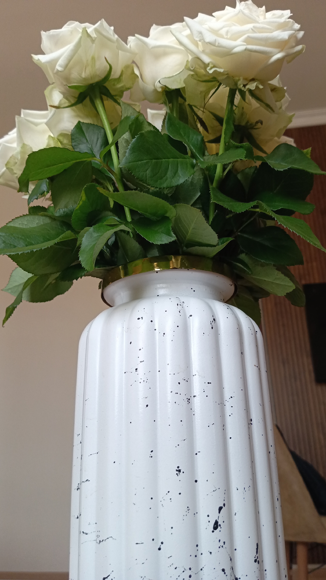 A close-up shot shows a bouquet of white roses with lush green leaves emerging from a white ribbed vase. The vase is speckled with black paint, giving it a distressed or artistic appearance. A thin gold band encircles the rim of the vase. The background is softly blurred, with a neutral-toned wall on the left and a suggestion of wooden paneling on the right. The lighting is natural and diffused, highlighting the textures of the flowers, leaves, and vase. No specific location in Ashtarak, Armenia, can be identified from this image.