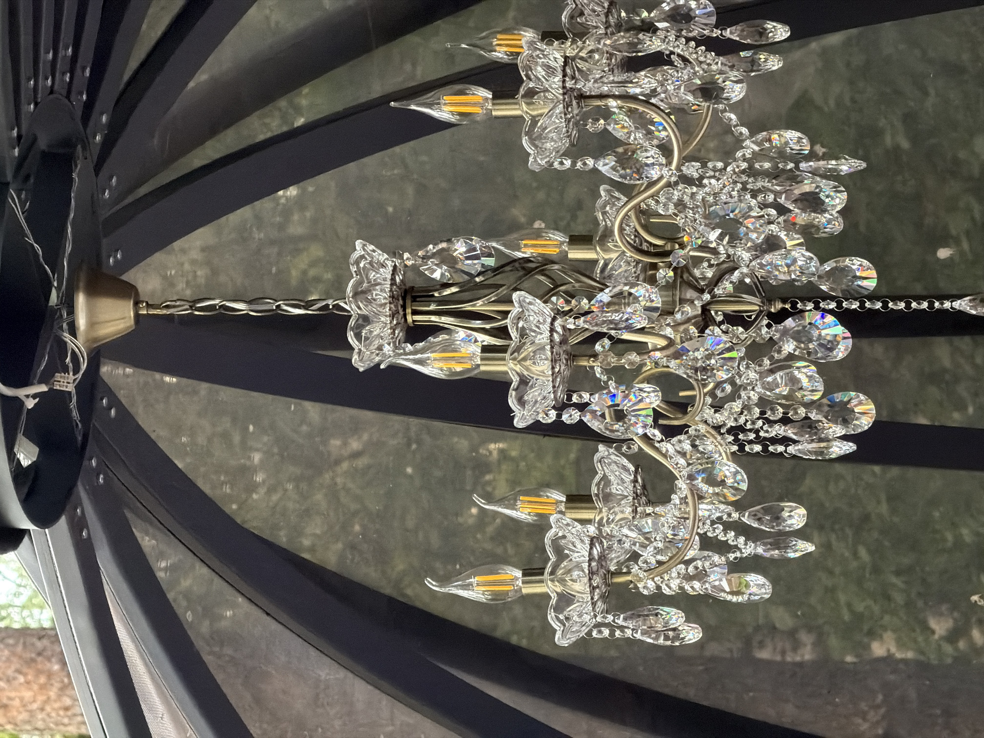 A brass chandelier with crystal accents hangs inside a dark, geodesic dome structure. The chandelier has multiple arms, each supporting a clear, flame-shaped LED bulb and adorned with tiered crystal cups and dangling faceted crystals that reflect rainbow colors. The background is a blurred view of green foliage, suggesting an outdoor setting. The dome's frame is visible as dark, curved beams. The chandelier is the primary subject, positioned centrally and well-lit. The overall impression is one of decorative illumination within a sheltered space. The location is specified as Rakitnitski selski Saveet, Belarus, though this information cannot be visually confirmed from the image itself.