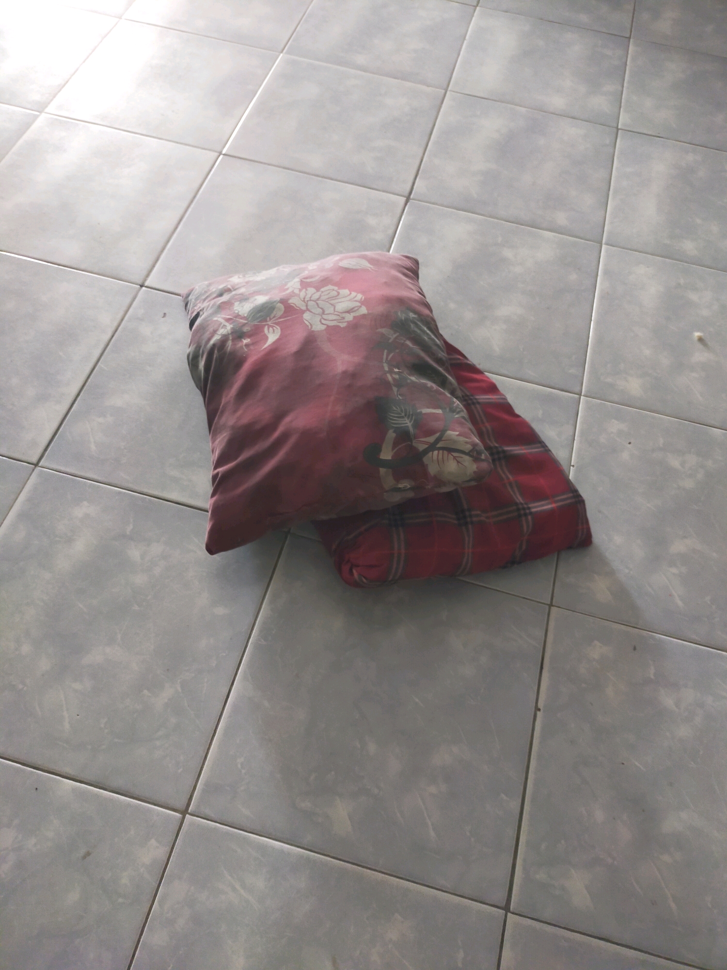 Two pillows are stacked on a tiled floor. The top pillow is red with a floral pattern. The bottom pillow is red with a plaid pattern. The floor is made of gray square tiles. The scene appears to be indoors, likely in a home. There is no information in the image to suggest the location is the city of Pekanbaru, Indonesia.
