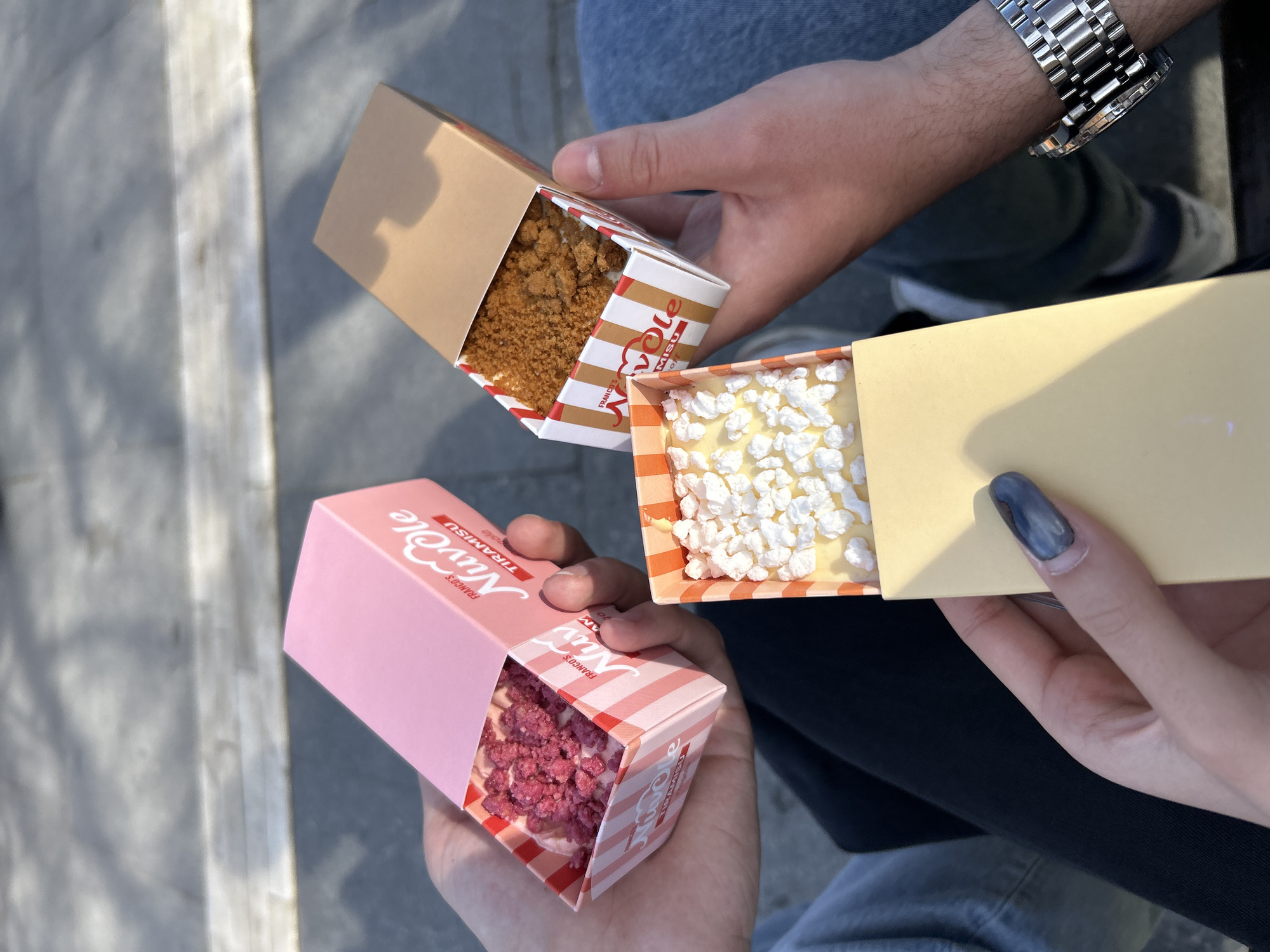 In the city of Sofia, Bulgaria, the image captures a candid moment with three individuals holding boxes of what appears to be tiramisu. The person on the right, presumably wearing a watch, holds a box filled with a crumbly, brown substance. The person on the left holds a box containing white, puffy objects. Finally, the person on the bottom holds a pink box containing small, red pieces. All boxes are of a similar rectangular shape and seem to be from the same brand. The people are seated. The background includes a concrete surface and blue denim fabric.