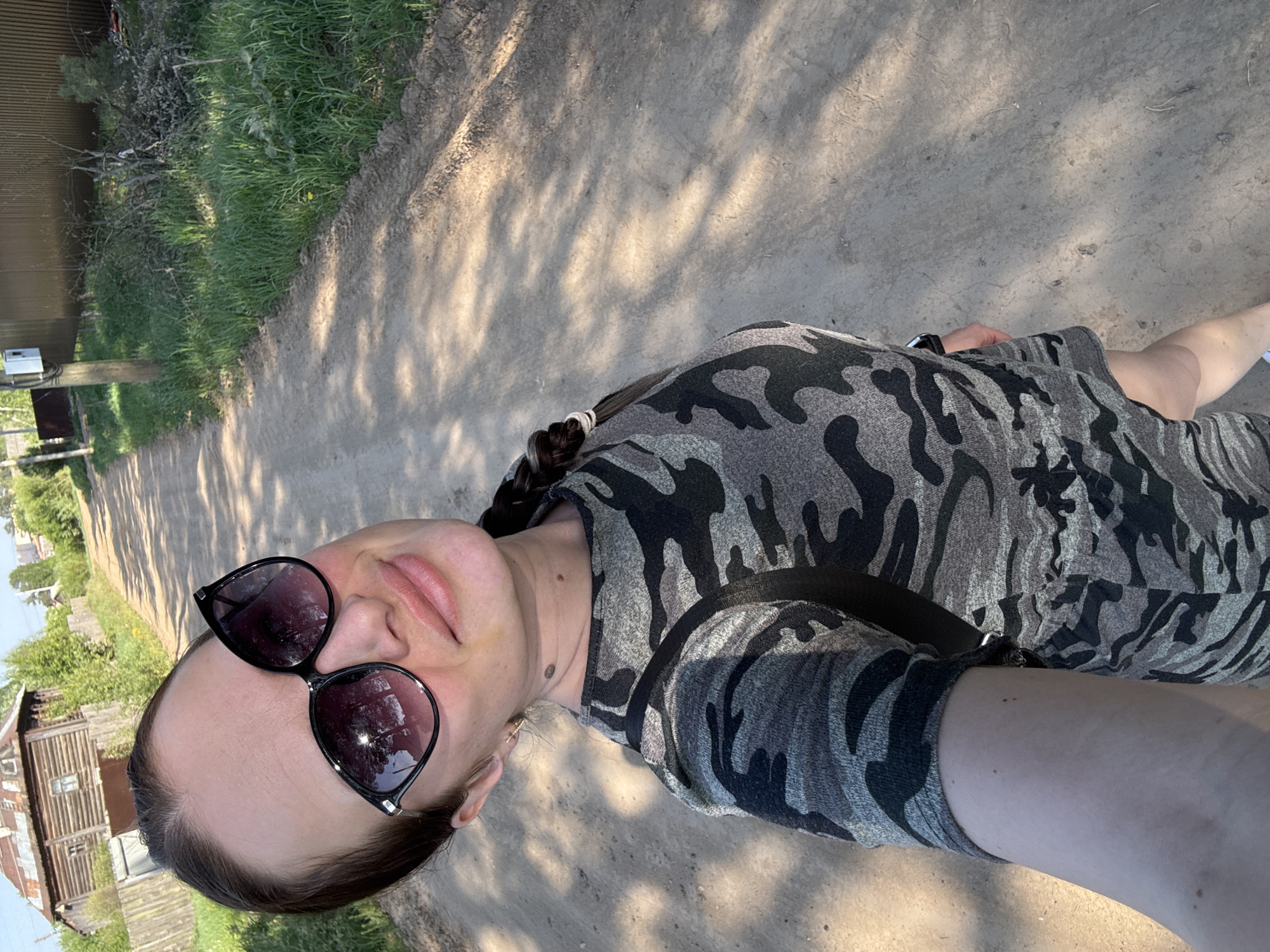 A woman with sunglasses and a camouflage pattern dress is standing on a dirt road. The location appears to be near the city of Rusalevka, Russia, as suggested by the rural environment with wooden houses and unpaved roads. She seems to be taking a selfie. The lighting suggests it is daytime. There is grass and some foliage beside the road, and shadows cast on the ground.