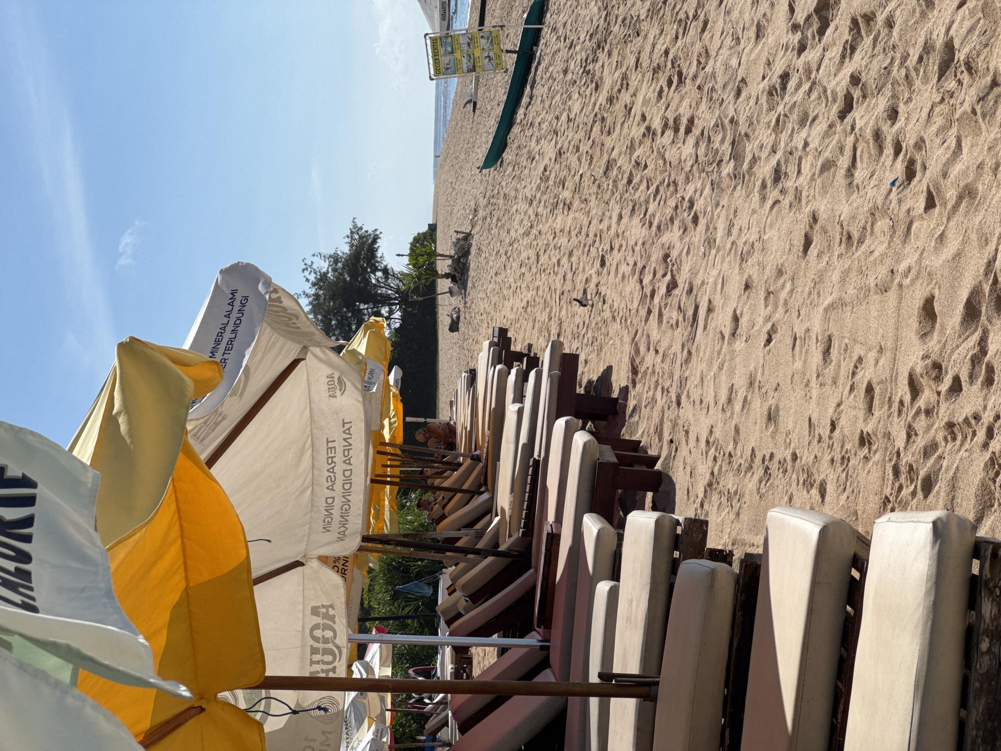 The image captures a sun-soaked scene at a beach in Unknown, Indonesia, where the warm-toned sand meets the blue of the sky. In the foreground, a row of lounge chairs, with weathered wooden frames and off-white cushions, lines the beach, providing a space for relaxation. Above the chairs, a cluster of beach umbrellas provides shade, their yellow and white canopies displaying advertisements for "AUGA" and "MINERAL ALAMI TERLINDUNGI," hinting at local products. Further down the beach, a sign that says "GEGER RENTAL" stands near a dark green canoe or kayak, suggesting water activities. The background shows a sparse tree line along the coast, and a few people can be seen scattered on the sand.