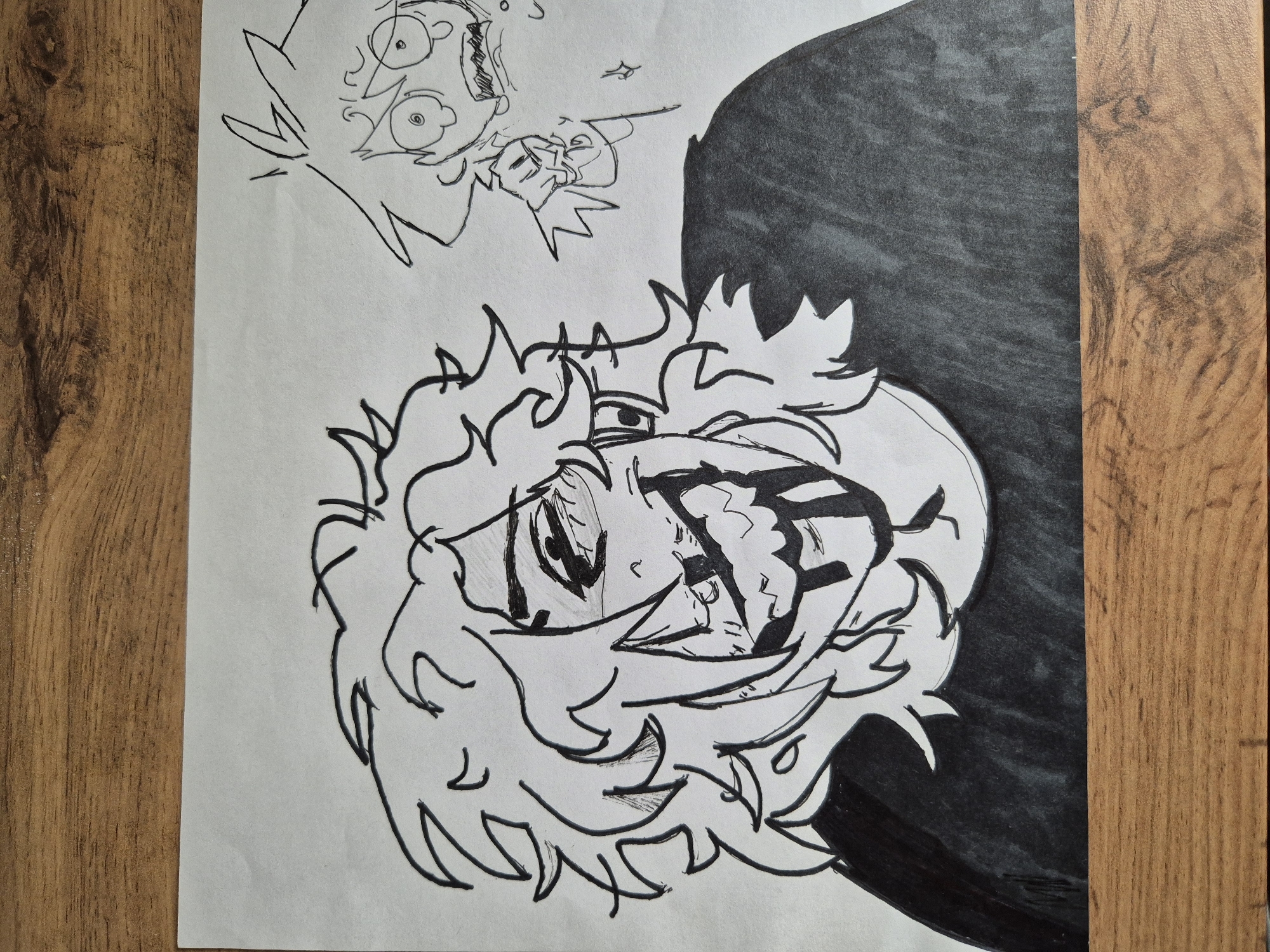 The image shows a black and white drawing on a sheet of paper placed on a wooden surface. The paper features two cartoon-style character drawings. The main drawing is of a person with spiky hair, large eyes, and a distressed expression, partially obscured by a black shaded area. Above it is a smaller, simpler cartoon depiction of another character with wide eyes and exaggerated features. The wooden surface appears to be a table or desk with a light brown, textured finish.