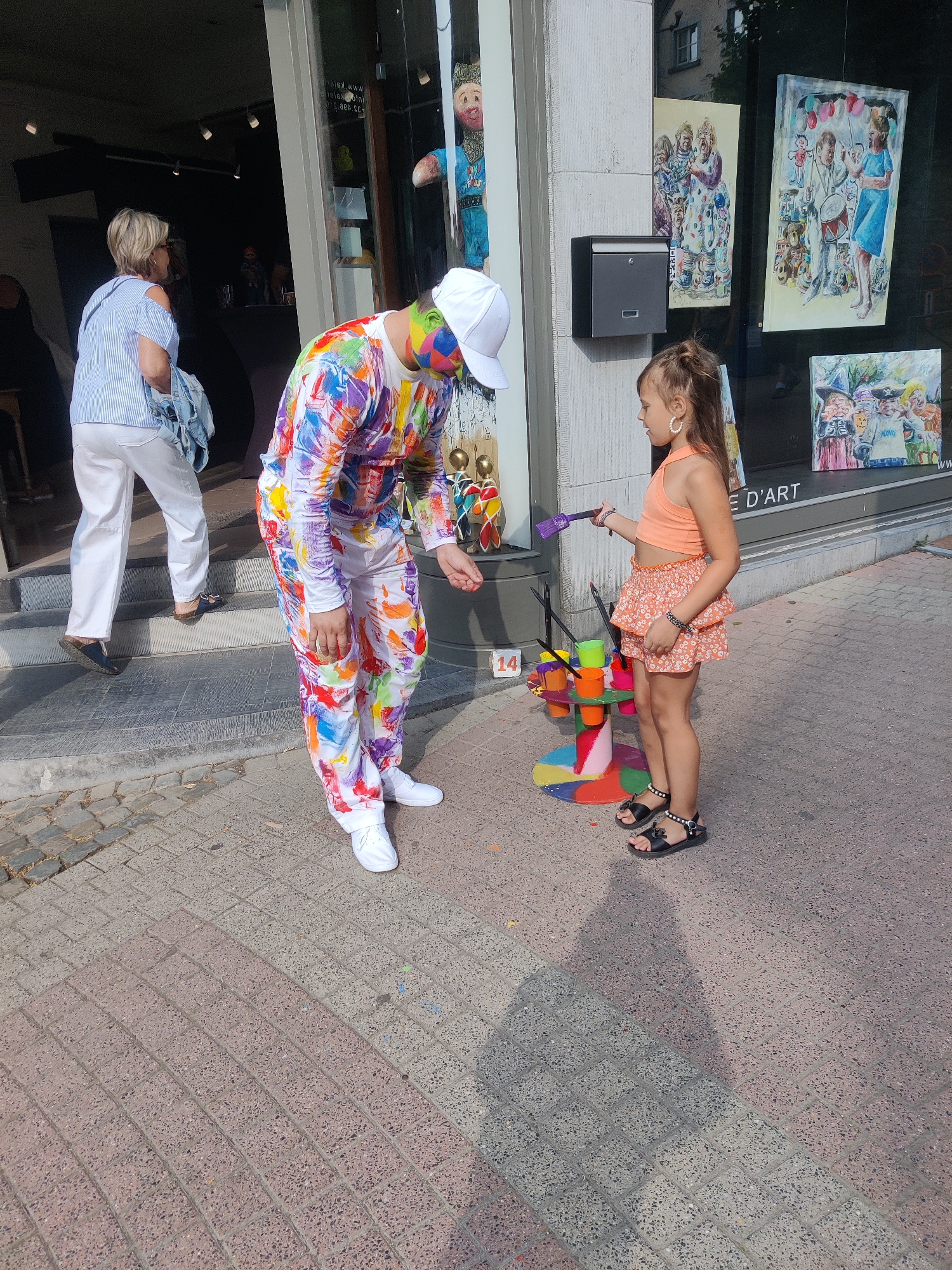 This image captures a vibrant street scene in Marche-en-Famenne, Belgium, likely on Rue des Casernes, given the artistic and cultural context suggested by the shop window displays. In the foreground, a young girl with brown hair, wearing a bright orange crop top and a floral skirt, stands on a paved sidewalk. She is holding a purple paintbrush in her right hand, facing a man dressed in a white hat and a full-body suit splattered with a multitude of colorful paint strokes. He appears to be interacting with the girl, possibly demonstrating a painting technique. To the left, a woman with blonde hair, wearing a blue and white striped shirt and white pants, walks away from the camera up a set of steps leading into a building. Behind the man and girl, a colorful display of art supplies and decorative elements is visible, including a tiered stand holding various colored paint pots and brushes. The shop window behind them showcases several framed paintings, depicting colorful and whimsical scenes, including what appears to be a puppet and other illustrated figures, suggesting the location is an art gallery or shop. The building itself has a modern facade with large glass windows. The ground is paved with a textured, patterned brick material. The sunlight casts shadows across the sidewalk, indicating it is daytime.