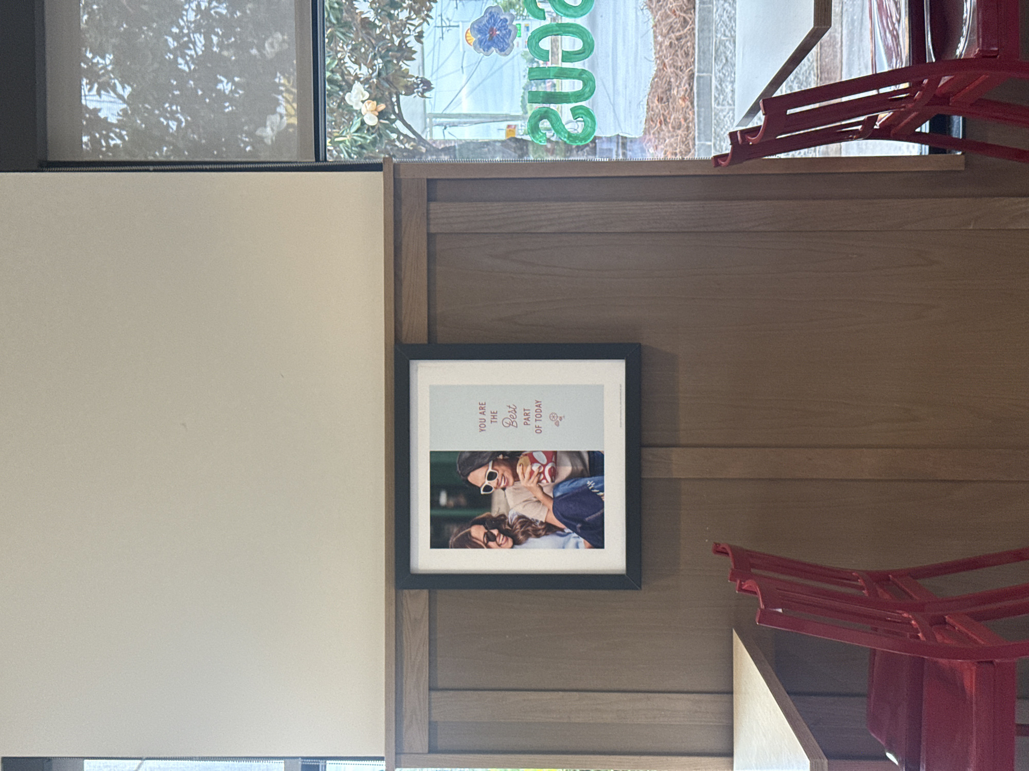 The image shows a framed picture hanging on a wooden paneled wall. The picture features two women, both wearing sunglasses, smiling and embracing. Above the picture is a window, through which a view of a building with a sign can be seen. Parts of red chairs are visible on the right side of the image. The picture includes the text "YOU ARE THE Best PART OF TODAY." The scene appears to be indoors. The location is in the city of Stone Mountain, United States of America.