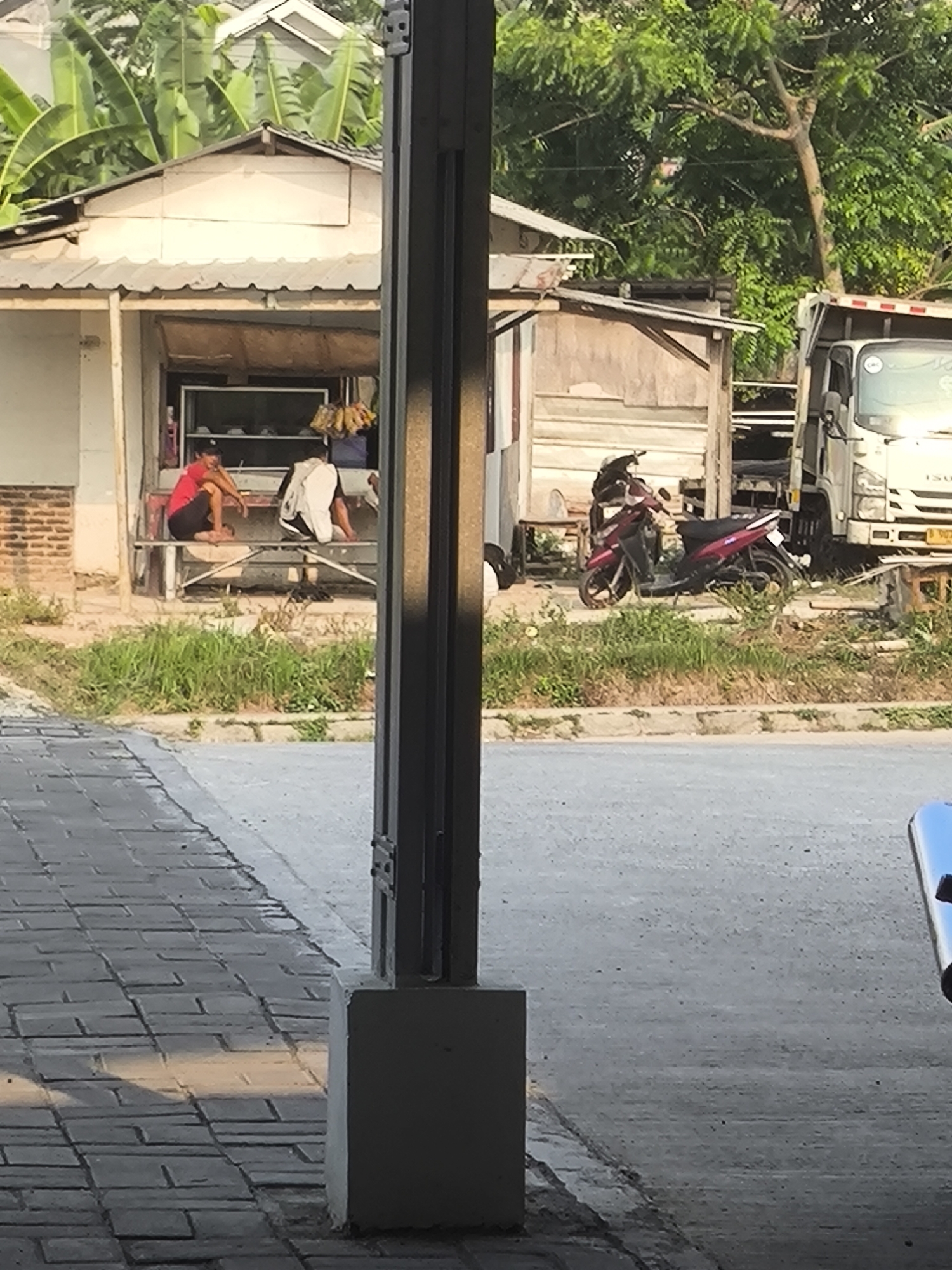 In Cilegon, Indonesia, an outdoor scene depicts a small open-air establishment with a white awning. In front of the establishment, two individuals are seated on a bench. One person, wearing a red shirt and black shorts, is looking to their left. The other individual, in a white shirt and dark shorts, is facing away from the viewer. Beside the establishment, a red motorcycle is parked. Further to the right, a white truck is visible, partially obscured by the establishment and a wooden structure. The ground in front of the establishment is covered in green grass, transitioning to a paved walkway composed of grey bricks in the foreground. The overall environment appears to be a roadside or market area.