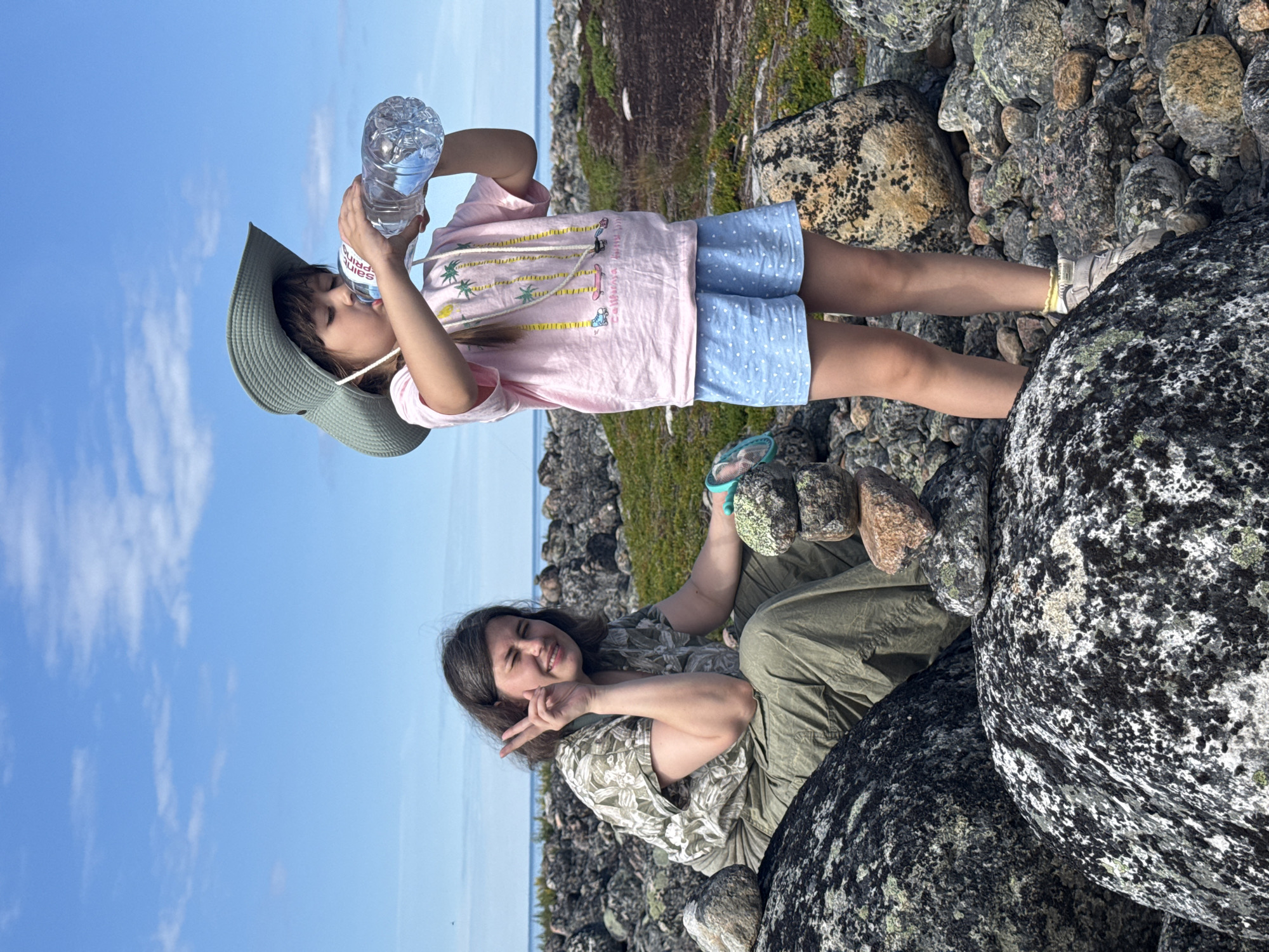 A woman and a young girl are outdoors on a rocky terrain with a body of water and blue sky in the background. The woman is seated on large boulders, wearing a green, floral-patterned short-sleeved shirt and olive-colored pants. She is making a peace sign with her right hand. The young girl stands to the right, wearing a green wide-brimmed hat, a pink t-shirt with palm trees and roller skates printed on it, and light blue shorts with white polka dots. She is drinking from a plastic bottle of water. In the foreground, various rocks are visible, with some stacked on top of each other. The environment appears to be a coastal or lakeside area with sparse vegetation.