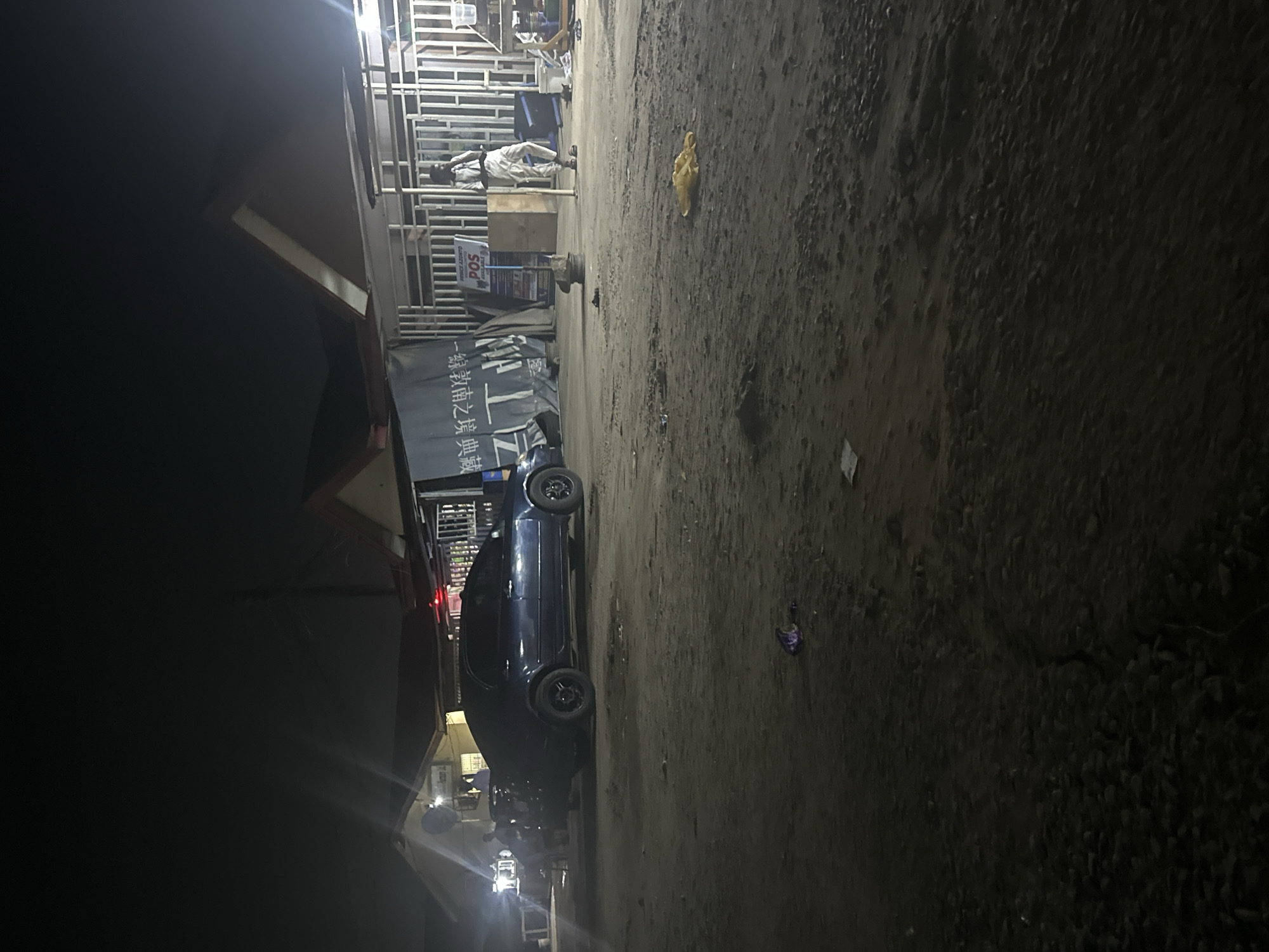 A nighttime scene in Abuja, Nigeria, shows a dark, unpaved area in front of a low-rise building. A blue sedan is parked on the left. A person in white clothing stands near a stall on the right, holding a mobile phone. A yellow cloth is on the ground in the middle of the scene. The building has a corrugated metal roof and an open-fronted structure. A large banner with Chinese characters hangs on the building. The ground is uneven and dusty, with scattered debris. Lights from the building illuminate the area.