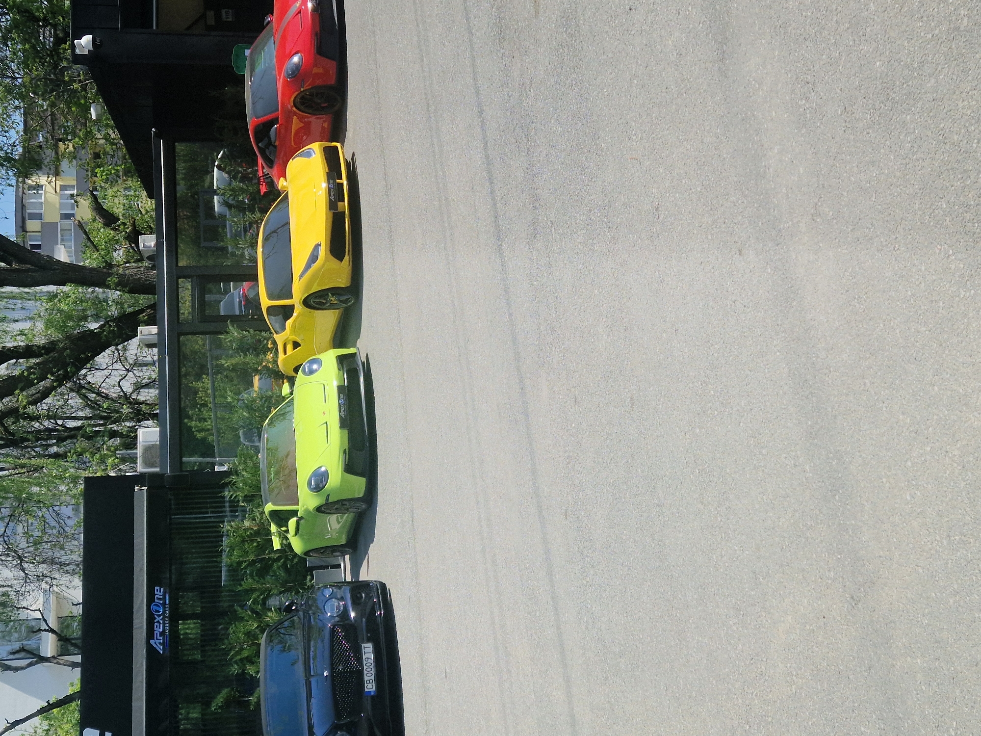 The image, rotated 90 degrees counter-clockwise, depicts a line of luxury and sports cars parked outdoors on a clear, sunny day. The vehicles are arranged along a light gray paved surface, likely a lot or driveway, adjacent to a modern building with dark paneling and large glass windows.
From what appears to be the top to the bottom of the rotated image, the cars include:
1. A red sports car, possibly a Porsche, visible at the top.
2. A vibrant yellow sports car, resembling a Lamborghini, positioned below the red car. It has "Apex One" visible on its front.
3. A lime green sports car, similar in style to a Porsche, located beneath the yellow car. This vehicle also bears "Apex One" branding on its front.
4. Partially visible at the very bottom, a black luxury car with a distinctive grille, likely a Bentley. Its license plate, "CB 0009 IT," is clearly legible.
The adjacent building features a prominent illuminated sign that reads "ApexOne LUXURY CARS," indicating the location is likely a dealership, showroom, or rental service specializing in high-end automobiles. Lush green trees frame the background and are reflected in the building's windows, adding to the bright, outdoor setting. No people are visible in the scene.
