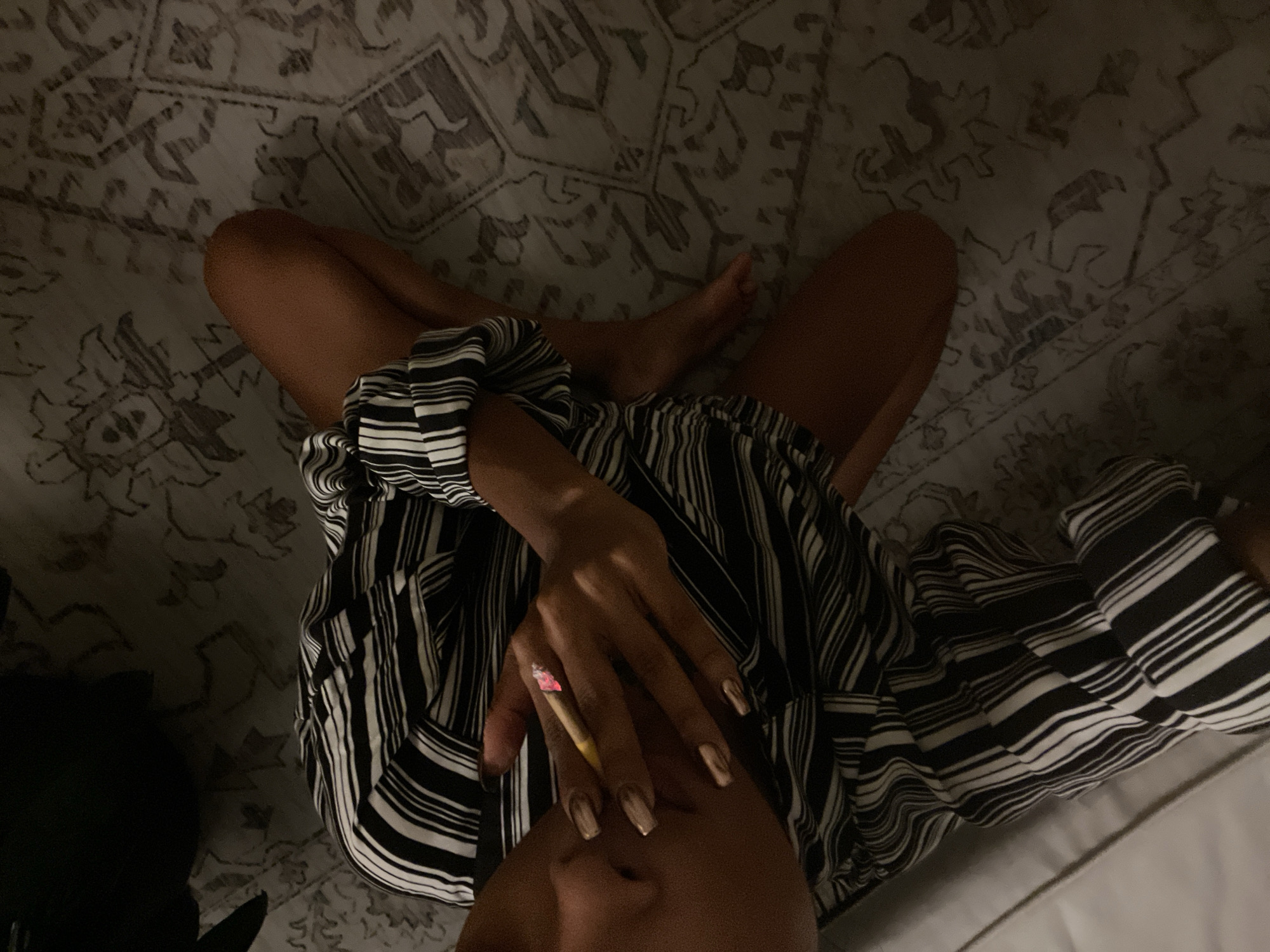 The image is a high-angle shot of a person sitting on a patterned rug, holding a lit cigarette. The person is wearing a black and white striped shirt with rolled-up sleeves and their legs are crossed. The cigarette is held between the index and middle fingers of the right hand, with the tip glowing red. The person's nails are painted a light metallic color. The background shows a sliver of a white couch and a dark object, possibly a bag. The lighting is dim, casting shadows on the rug. The image does not provide any information about the location of Singapore, Singapore.