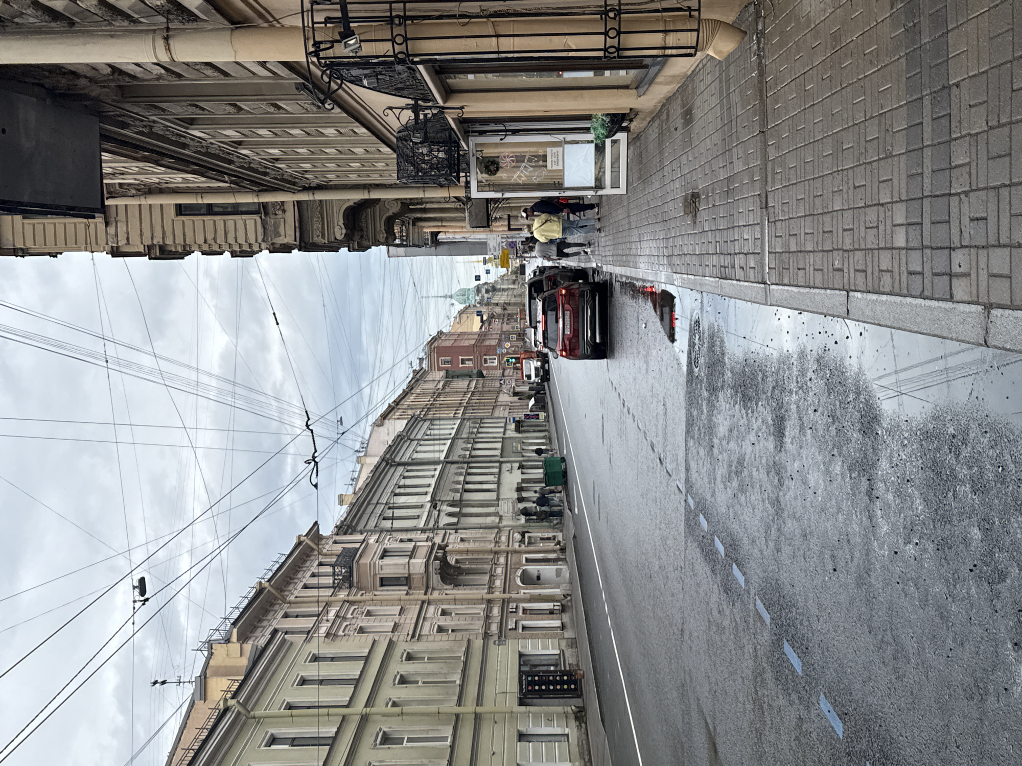 The image depicts a wet street scene in Saint Petersburg, Russia, with overcast skies. The perspective is from a sidewalk looking down a street lined with multi-story buildings on both sides. On the left, the buildings are a pale yellow and light beige, with numerous windows and decorative architectural details. A red car is parked on the wet asphalt road, which has visible white and blue lane markings. Several people are walking on the sidewalk on the right side of the street. The buildings on the right are a light beige color, with some showing shopfronts and ornate balconies. Overhead, a complex network of wires is visible against the sky. A domed building's spire is visible in the distance down the street. The overall impression is that of a historic European city on a rainy or recently rained-on day.