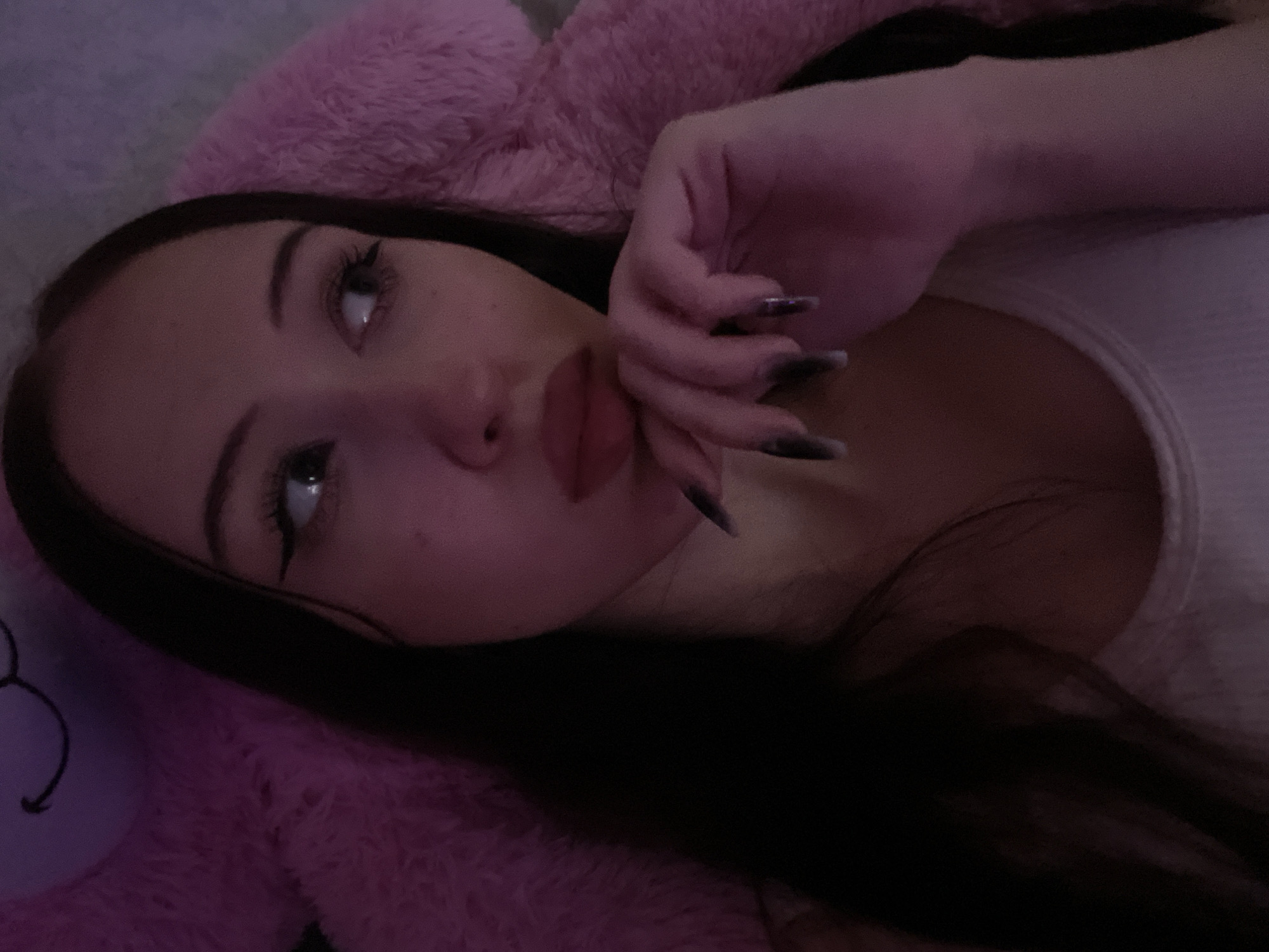 The image shows a close-up, low-angle shot of a young woman lying down, possibly on a large pink stuffed animal. She has long dark hair, dark eyeliner, and dark lipstick. Her right hand is resting on her chin, with long, dark fingernails. The lighting is dim and pinkish. There is no visible indication of the city of Votkinsk, Russia.