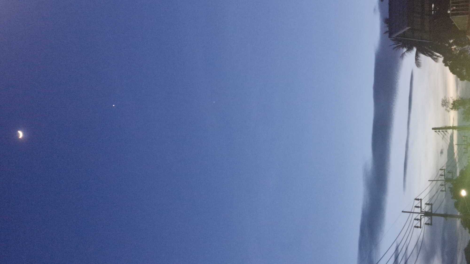This image depicts a dusky sky above the city of Ban Bang Rak, Thailand. A crescent moon is visible in the upper left quadrant, accompanied by a small, bright star below and to its right. In the lower portion of the frame, power lines stretch across the scene, supported by utility poles. A distant house with a dark roof and palm trees are visible on the right, and faint clouds are present near the horizon, suggesting the twilight hour. The overall scene is tranquil and observational.